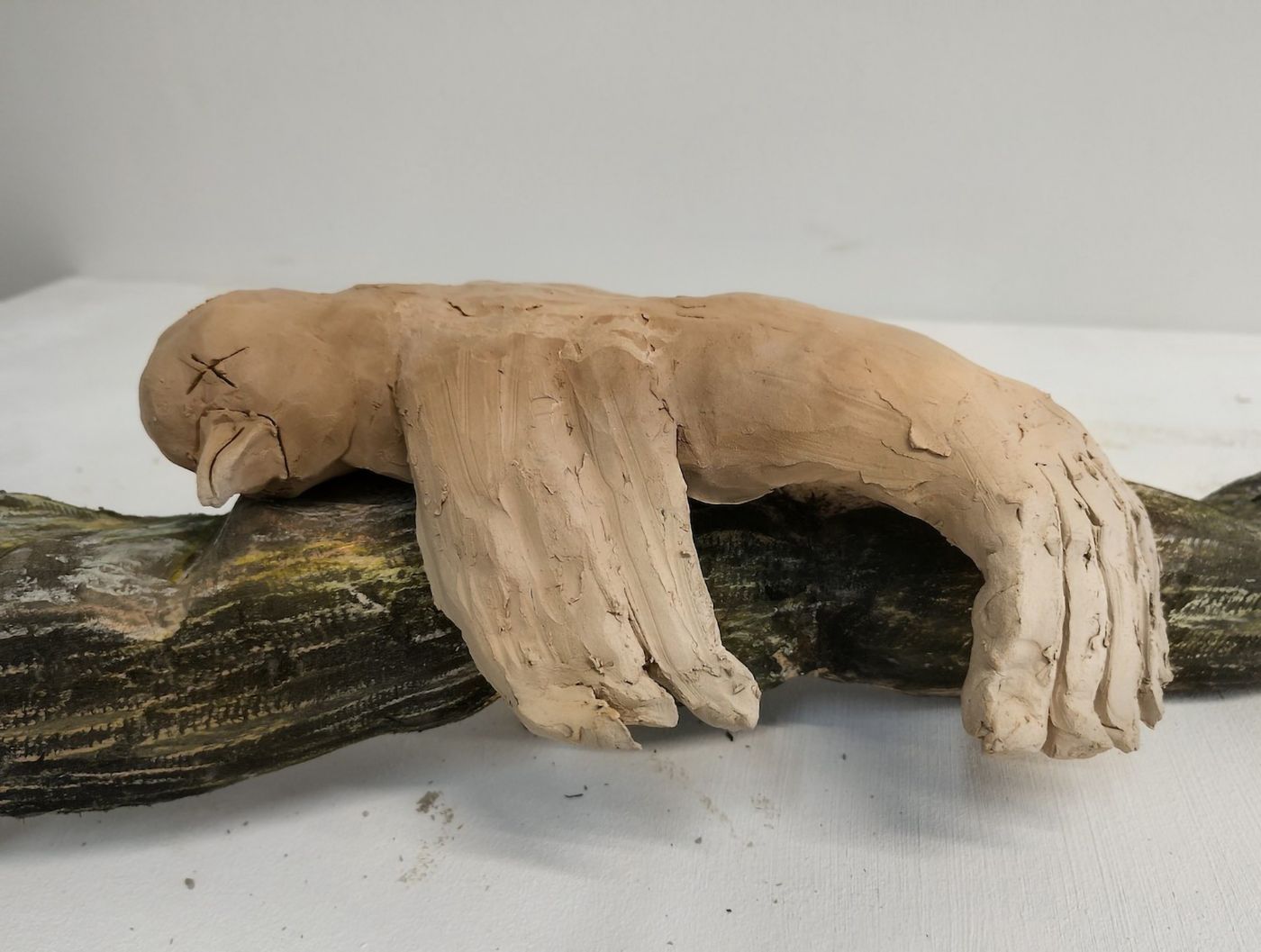 Eine aus Ton geformte Faultierfigur ruht auf einem bemalten Ast aus Holz auf einem Tisch
