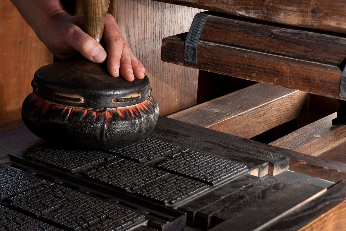 A hand uses a large leather printing tool on a wooden printing press with set type
