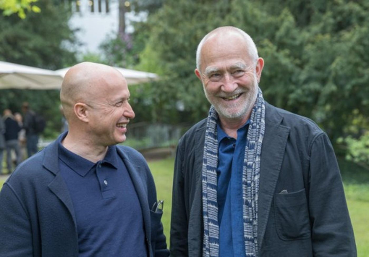 Zwei Männer lachen entspannt in einem grünen Garten, umgeben von Bäumen und Gästen