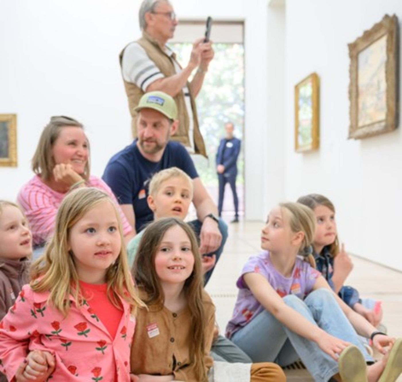 Eine Gruppe von Kindern und Erwachsenen sitzt fröhlich in einer Kunstgalerie.