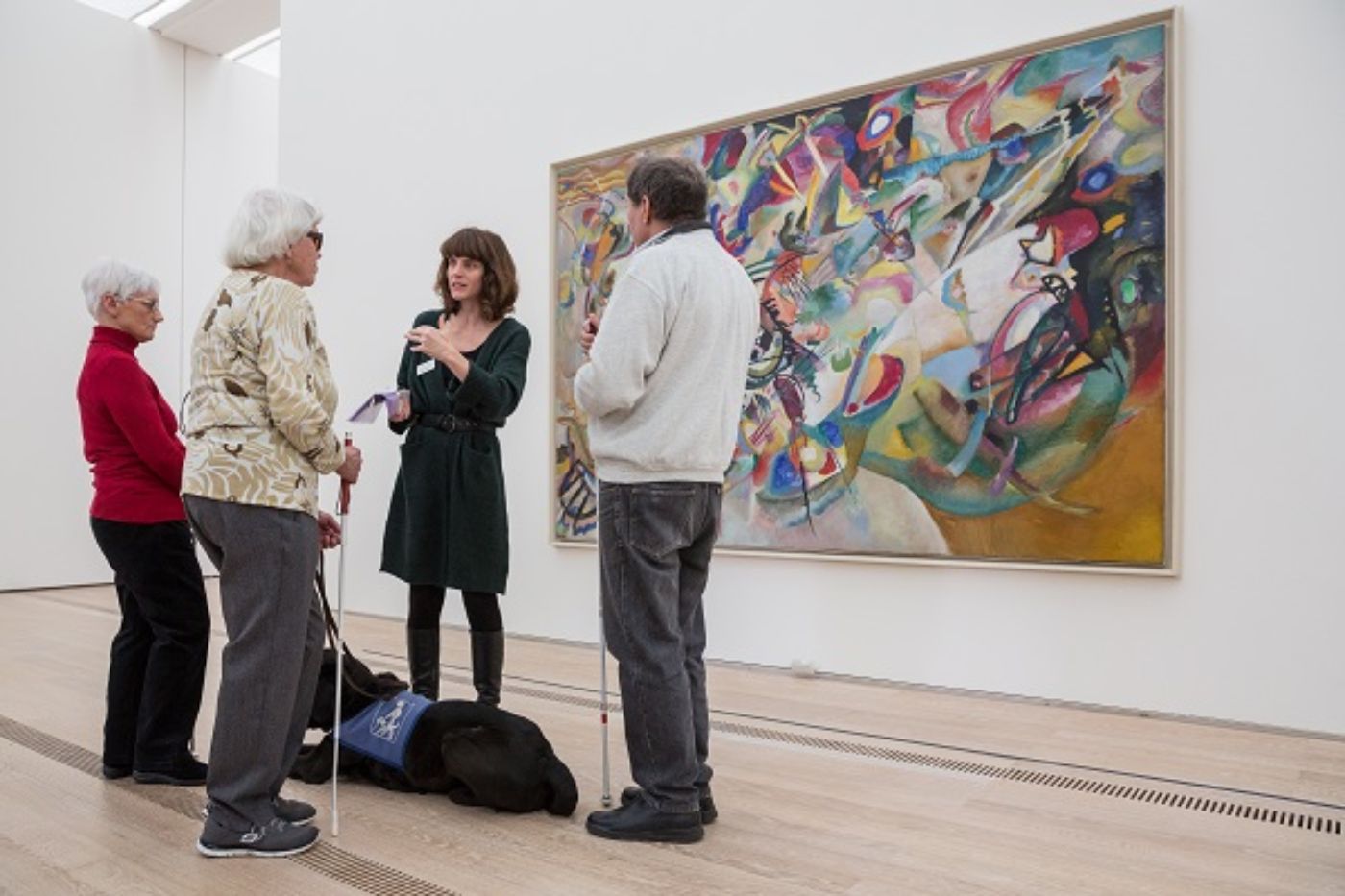 Eine Frau führt drei Personen mit Blindenstöcken vor einem bunten Gemälde in einer Galerie