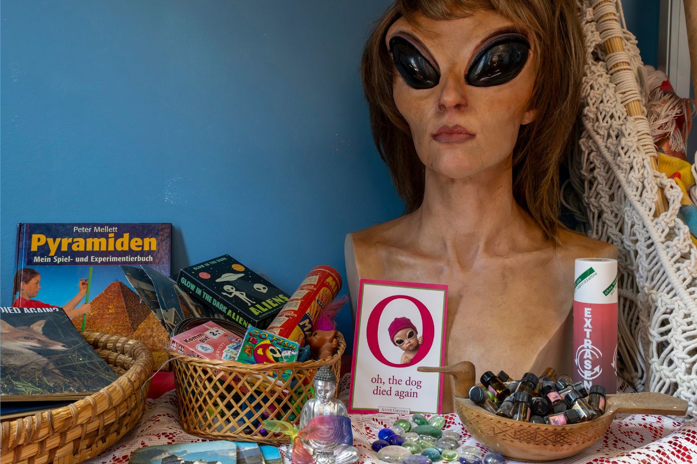 Ein Regal zeigt Pyramidenbücher, Alien-Spielzeug, und eine Statue mit grossen, dunklen Augen.