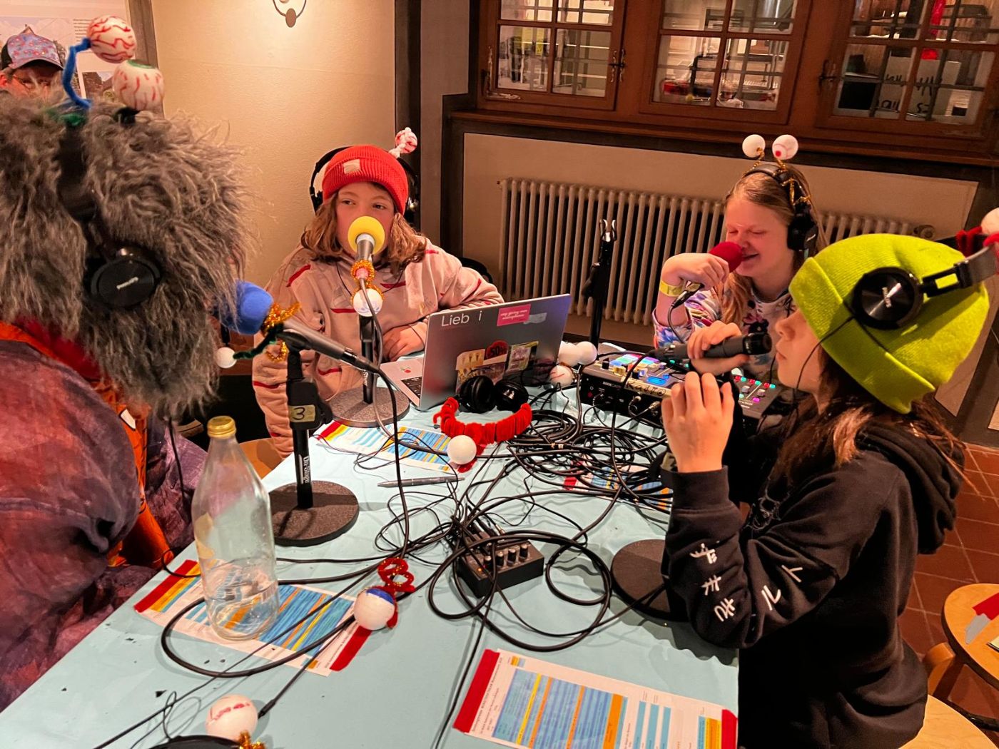 Drei Kinder mit Kopfhörern und Mikrofonen sitzen um einen Tisch voller Radiotechnik