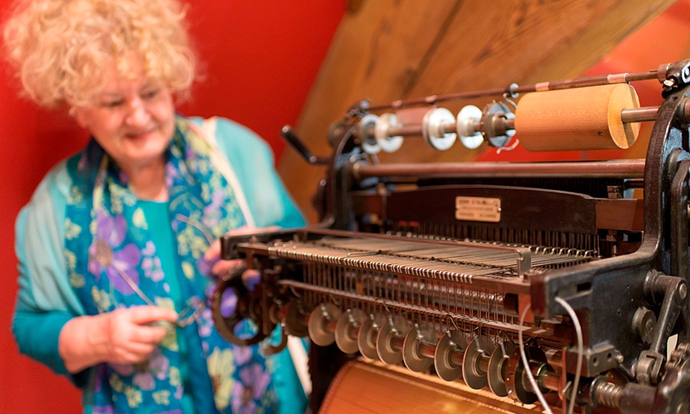 Eine Frau betrachtet interessiert eine alte Webmaschine mit roten Wänden im Hintergrund