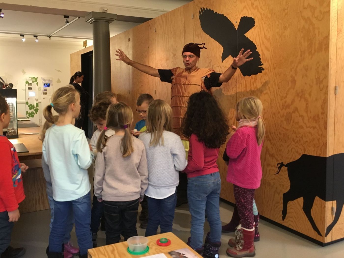 Zehn Kinder umringen einen Erzähler in Tierkostüm vor Holzwand mit Tierillustrationen