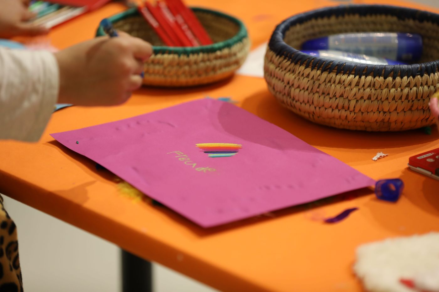Auf einem orangen Tisch liegen Malutensilien und ein pinkes Blatt Papier, auf dem eine Kinderhand malt.