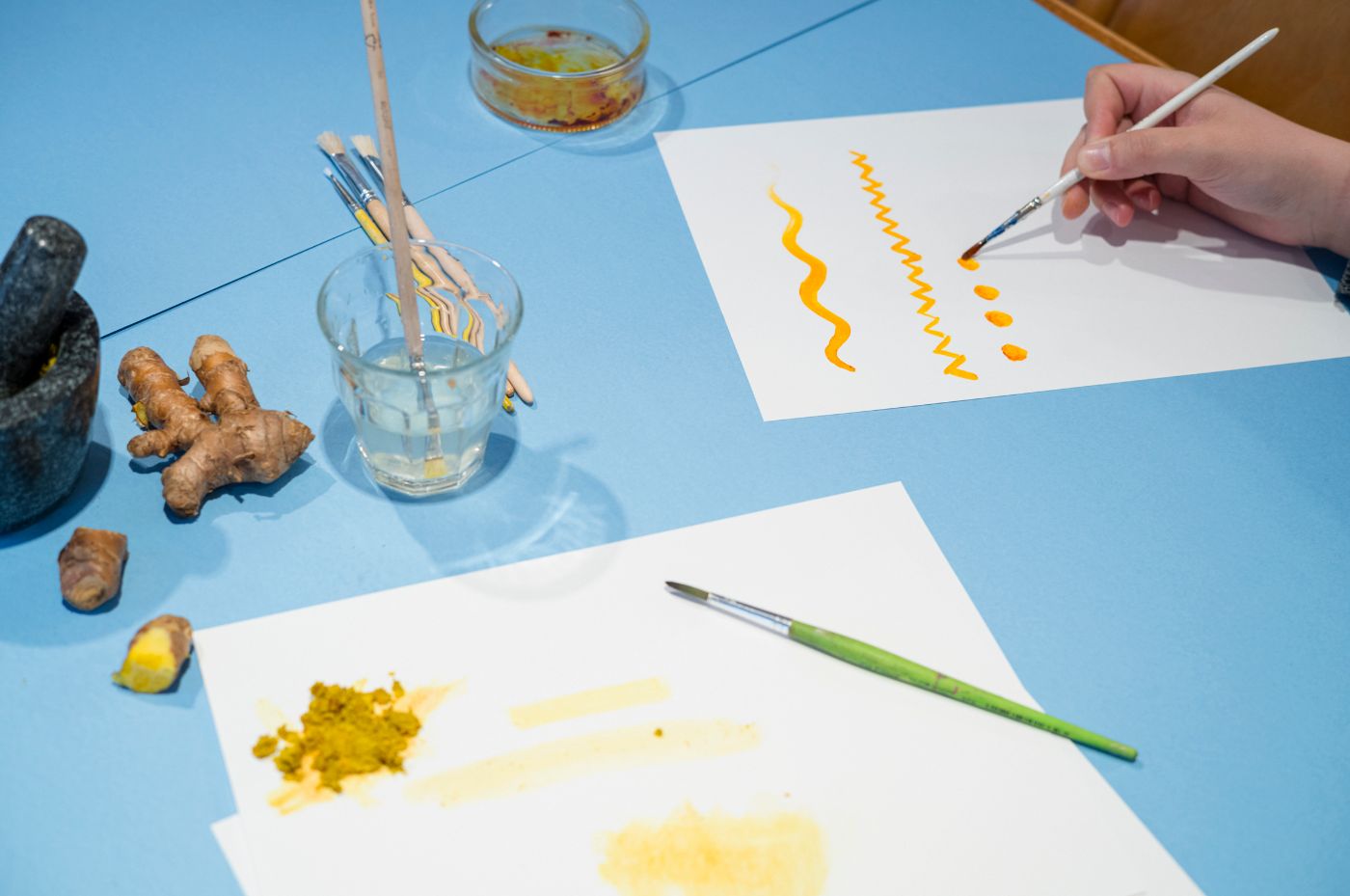 Auf einem hellblauen Tisch liegen Blätter, auf denen mit gelber Farbe gemalt wurde und gerade auch wird.