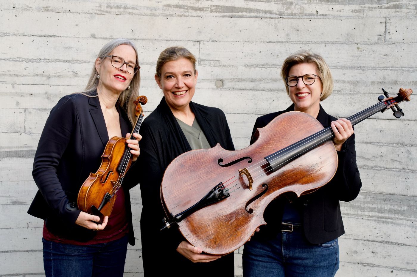 Drei Frauen stehen lächelnd vor einer Betonwand, halten Geige, Cello und Kontrabass in den Händen