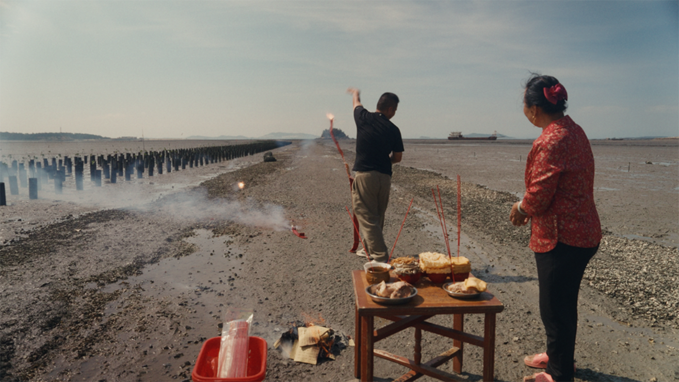 Zwei Personen führen ein Ritual am Strand durch, mit Essen und Räucherstäbchen auf einem Tisch