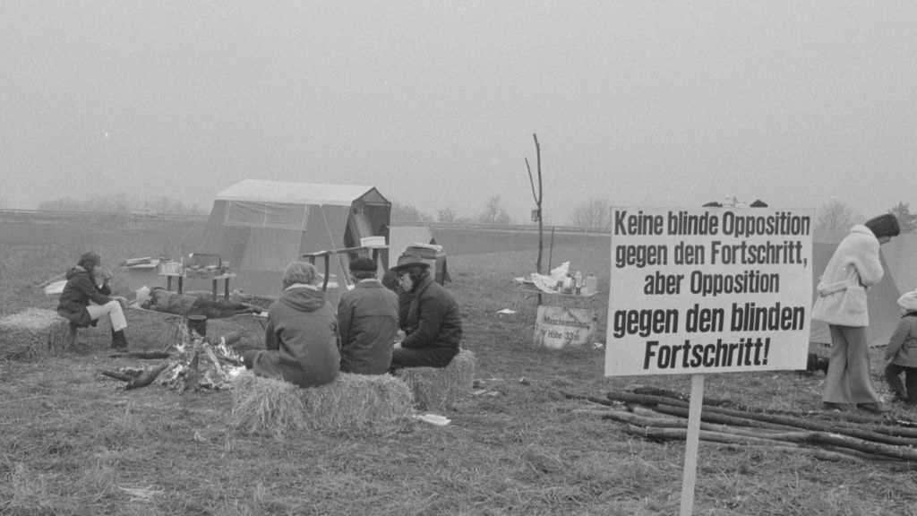 Eine Gruppe von Menschen sitzt auf Strohballen um ein Lagerfeuer; ein Zelt steht im Hintergrund.