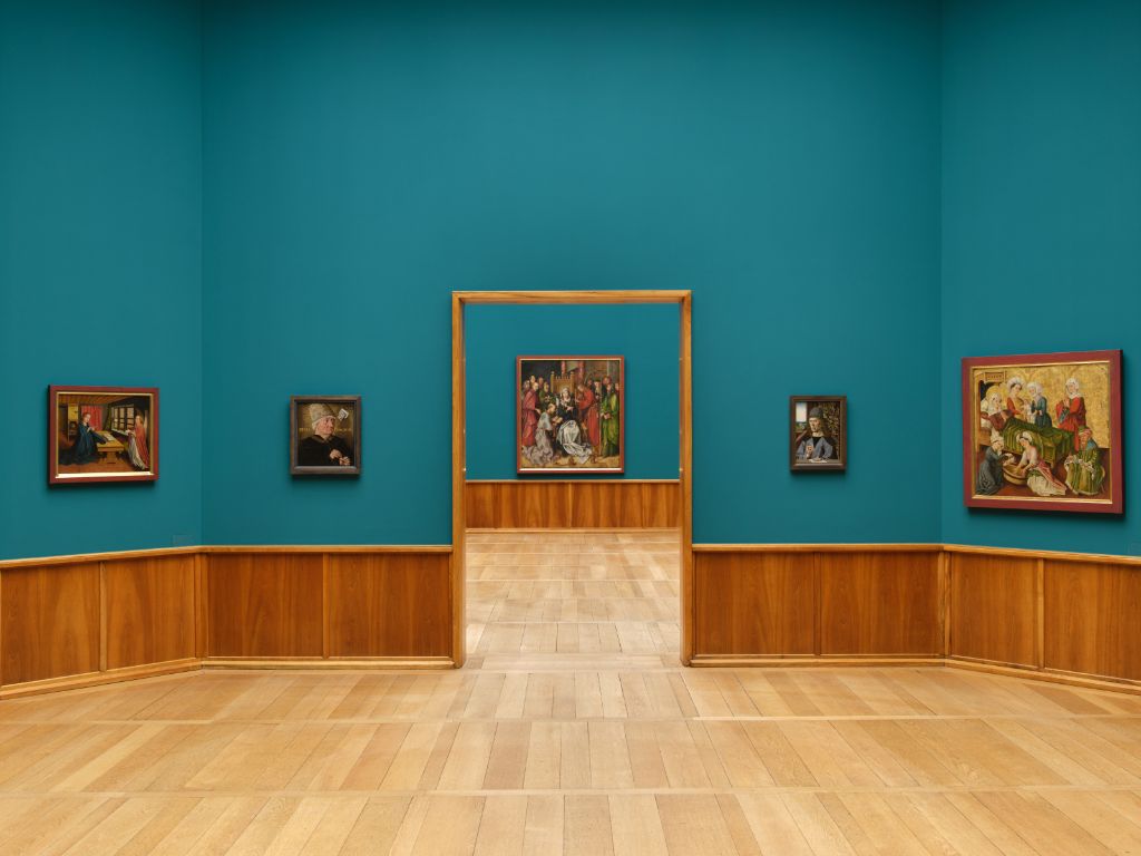 A bright exhibition room displays paintings and a sculpture on shiny wooden floors