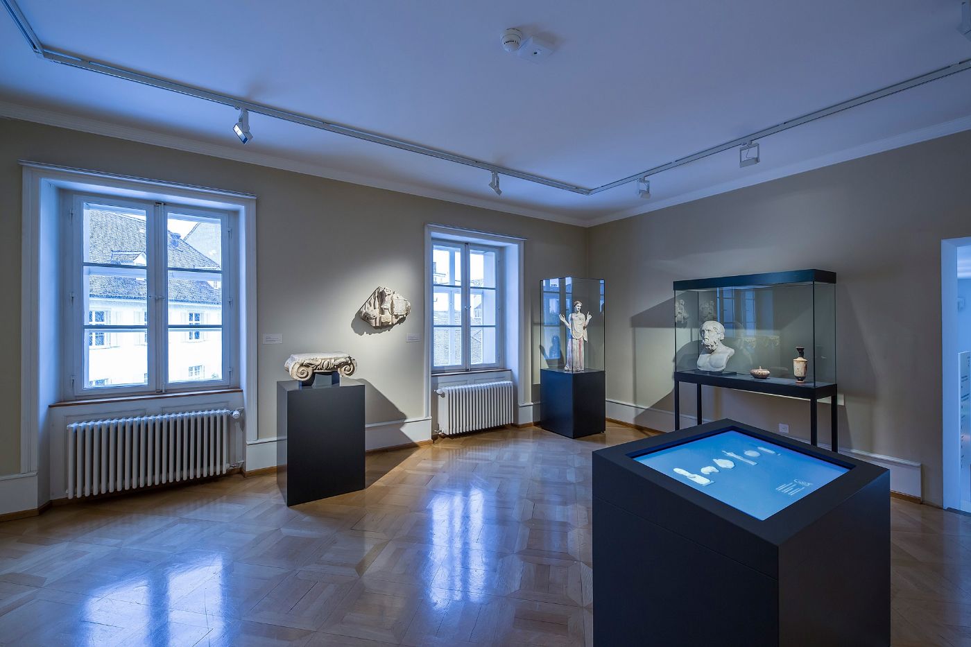 Ein ruhiger Museumssaal mit antiken Büsten und Statuen in Vitrinen, Licht strömt herein