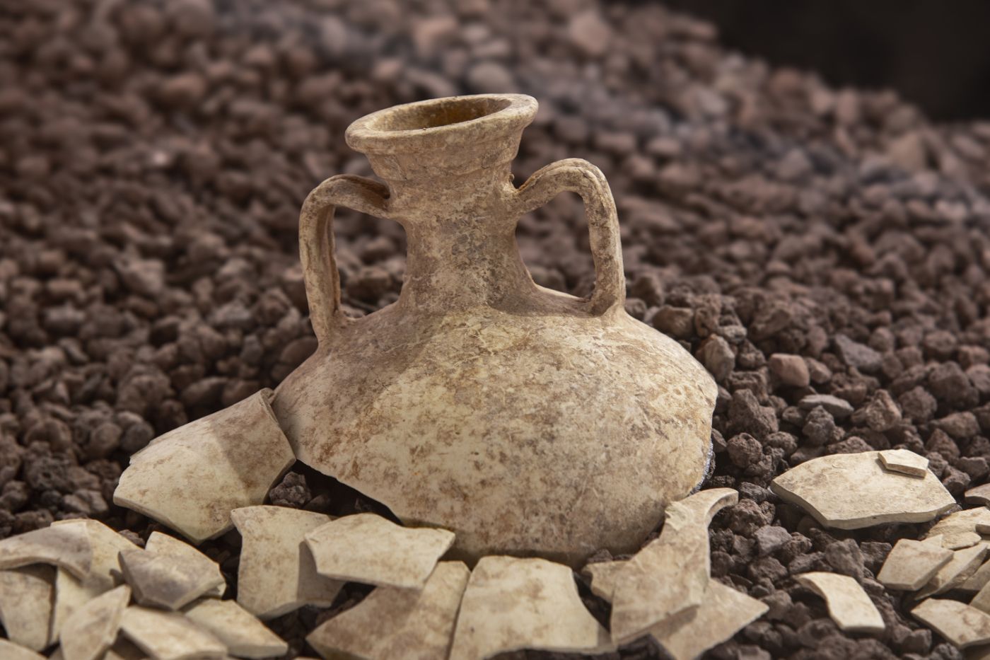 An ancient, broken clay jug rests on a ground of dark stones