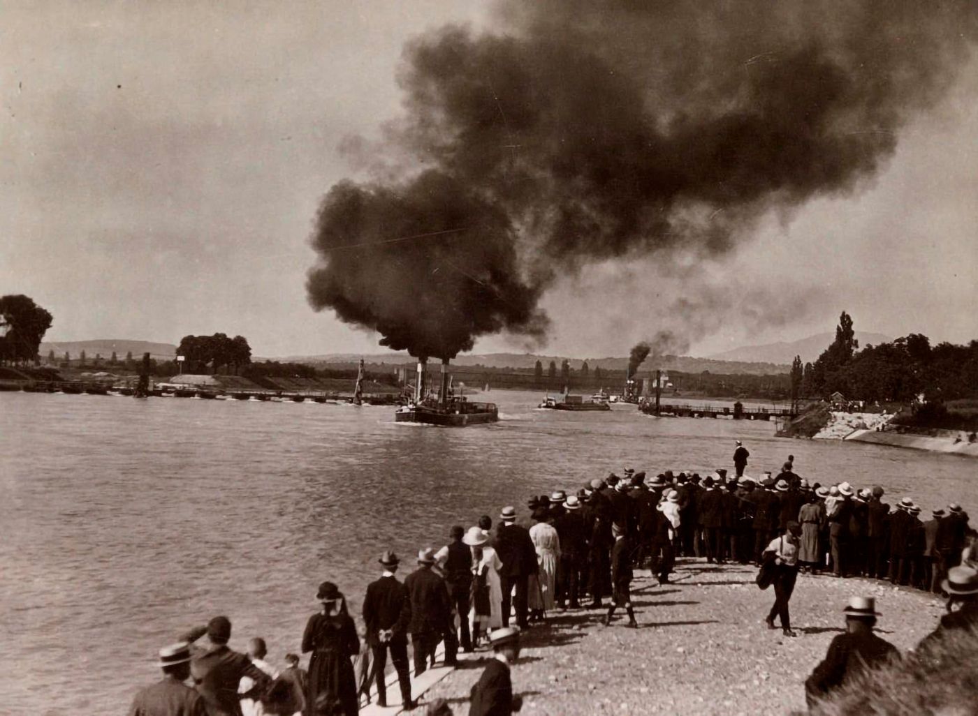 Une foule regarde des bateaux à vapeur avec des cheminées fumantes naviguer sur le fleuve
