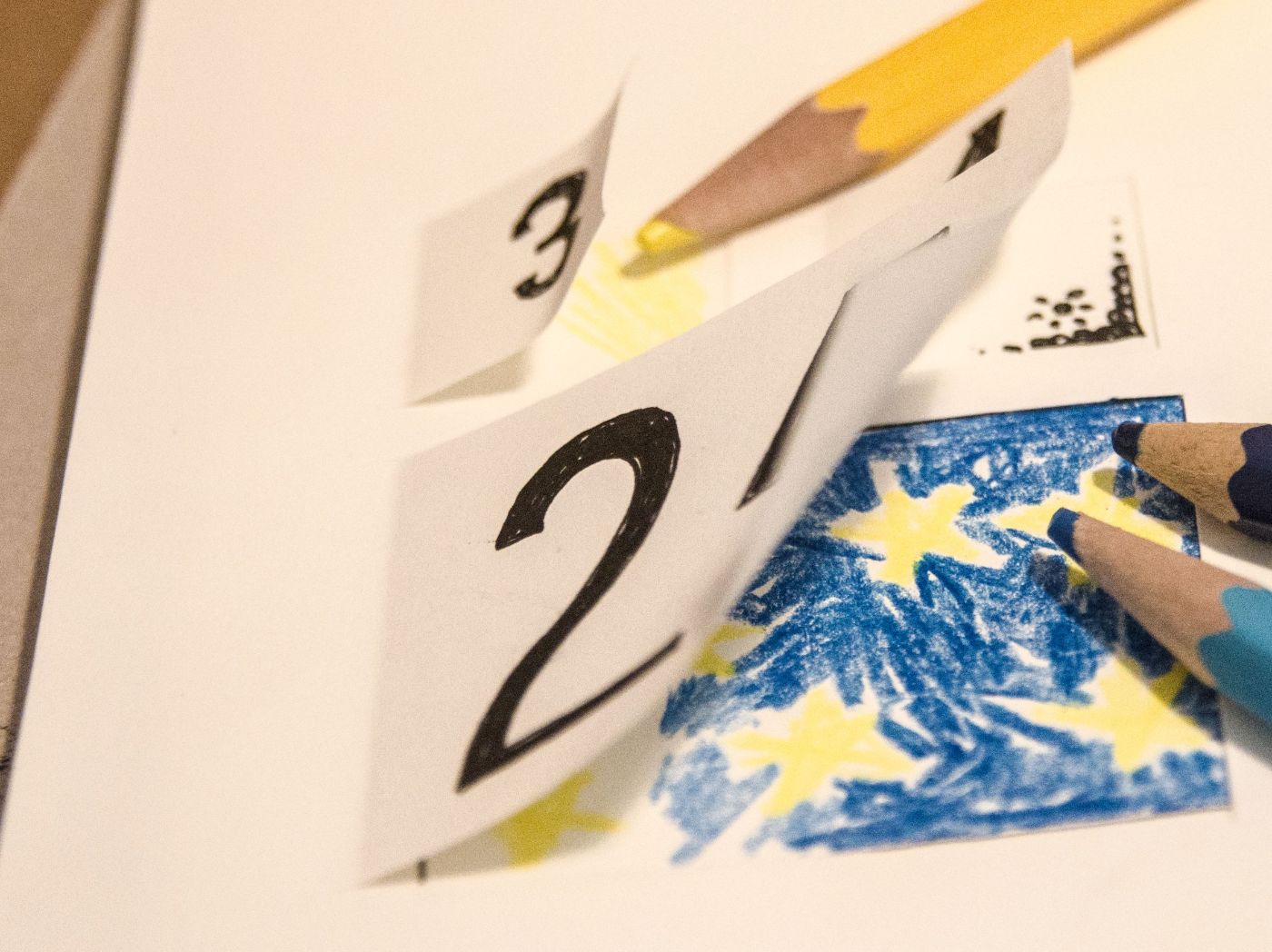 Auf einem Tisch liegen Buntstifte und ein Adventskalender mit bunten Zeichnungen und Zahlen.