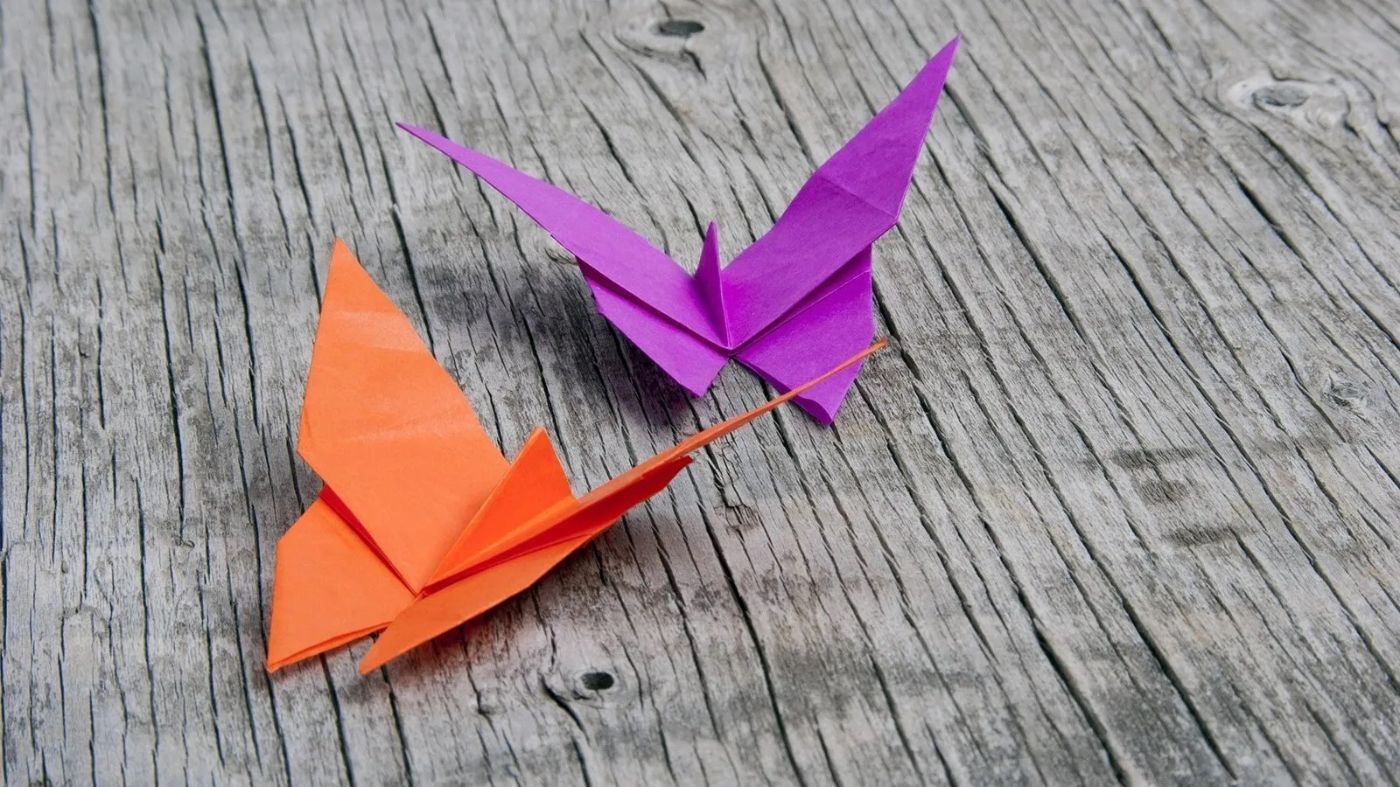 Zwei bunte Origami-Schmetterlinge in Orange und Lila ruhen auf einer verwitterten Holzoberfläche