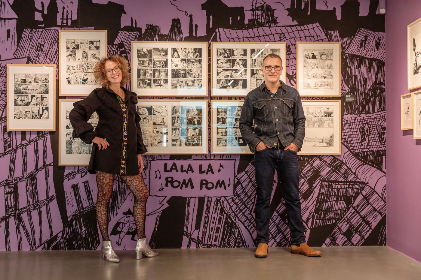 Zwei Personen stehen vor einer Wand mit gerahmten Comiczeichnungen und lilafarbenem Hintergrund.