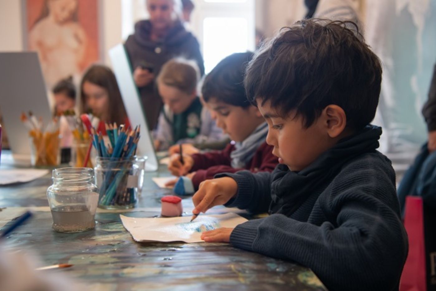 Ein Kind malt konzentriert auf Papier, umgeben von Malutensilien und weiteren Kindern