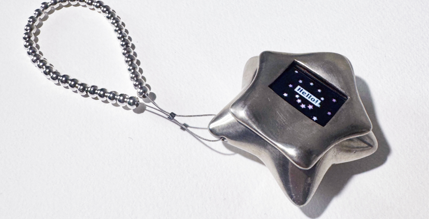 Ein silberner Sternanhänger mit digitalem Display zeigt "Hello!" und baumelt an einer Perlenkette