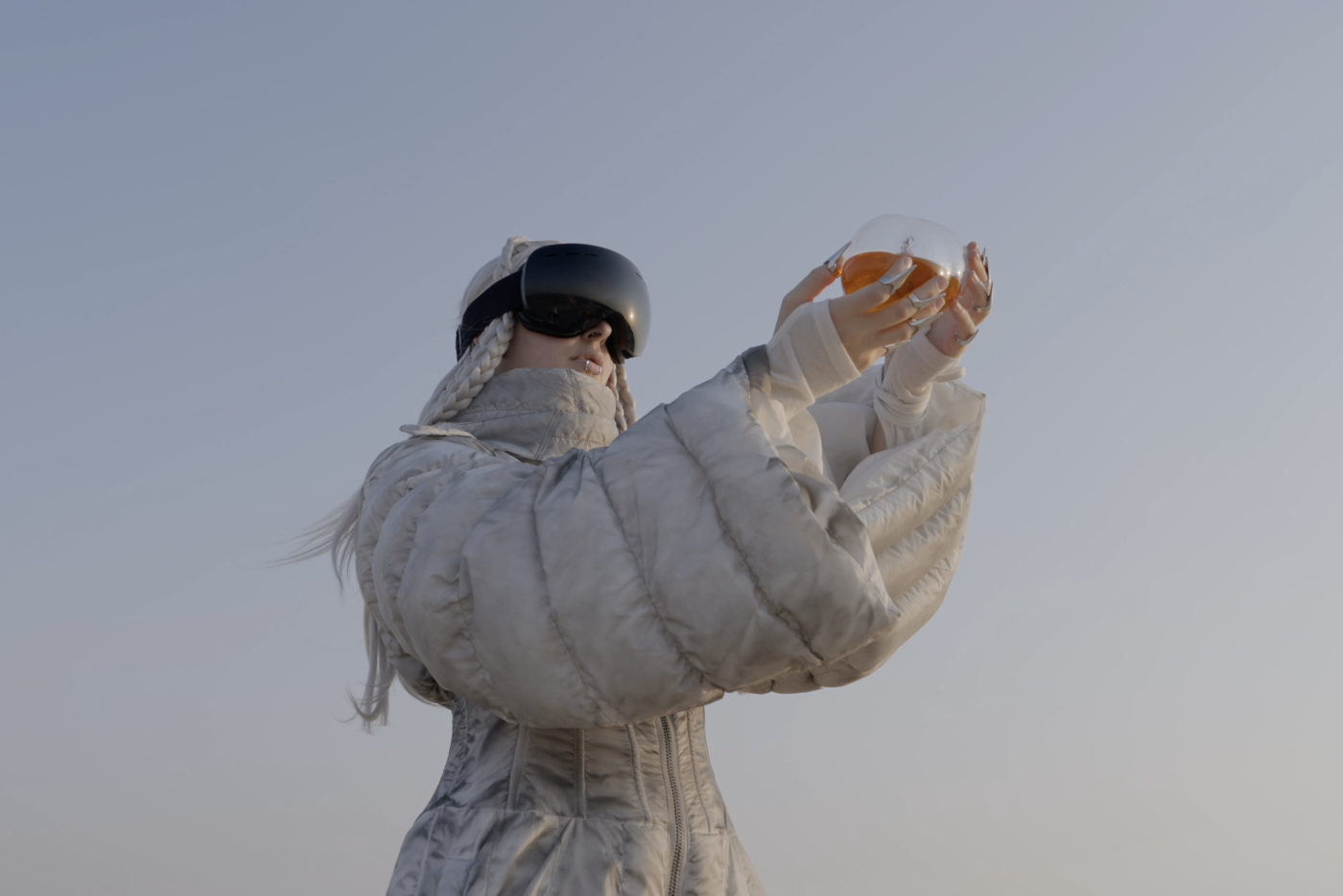 Eine Person in gepolsterter Kleidung und VR-Brille hält ein leuchtendes Gefäss gen Himmel