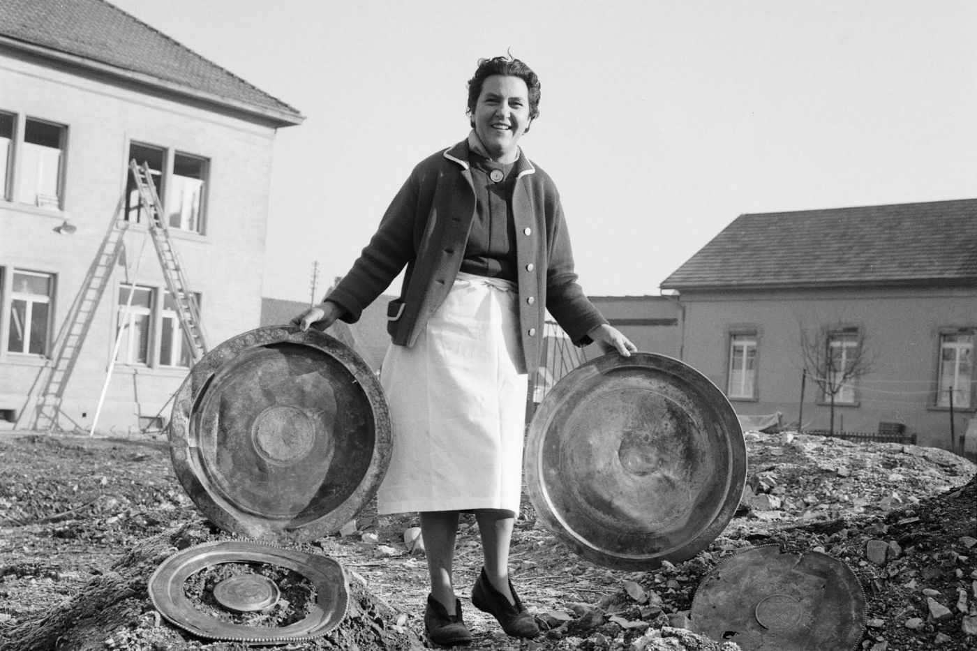 Eine Frau hält drei grosse, runde Metallschalen in einem offenen Gelände vor Gebäuden.