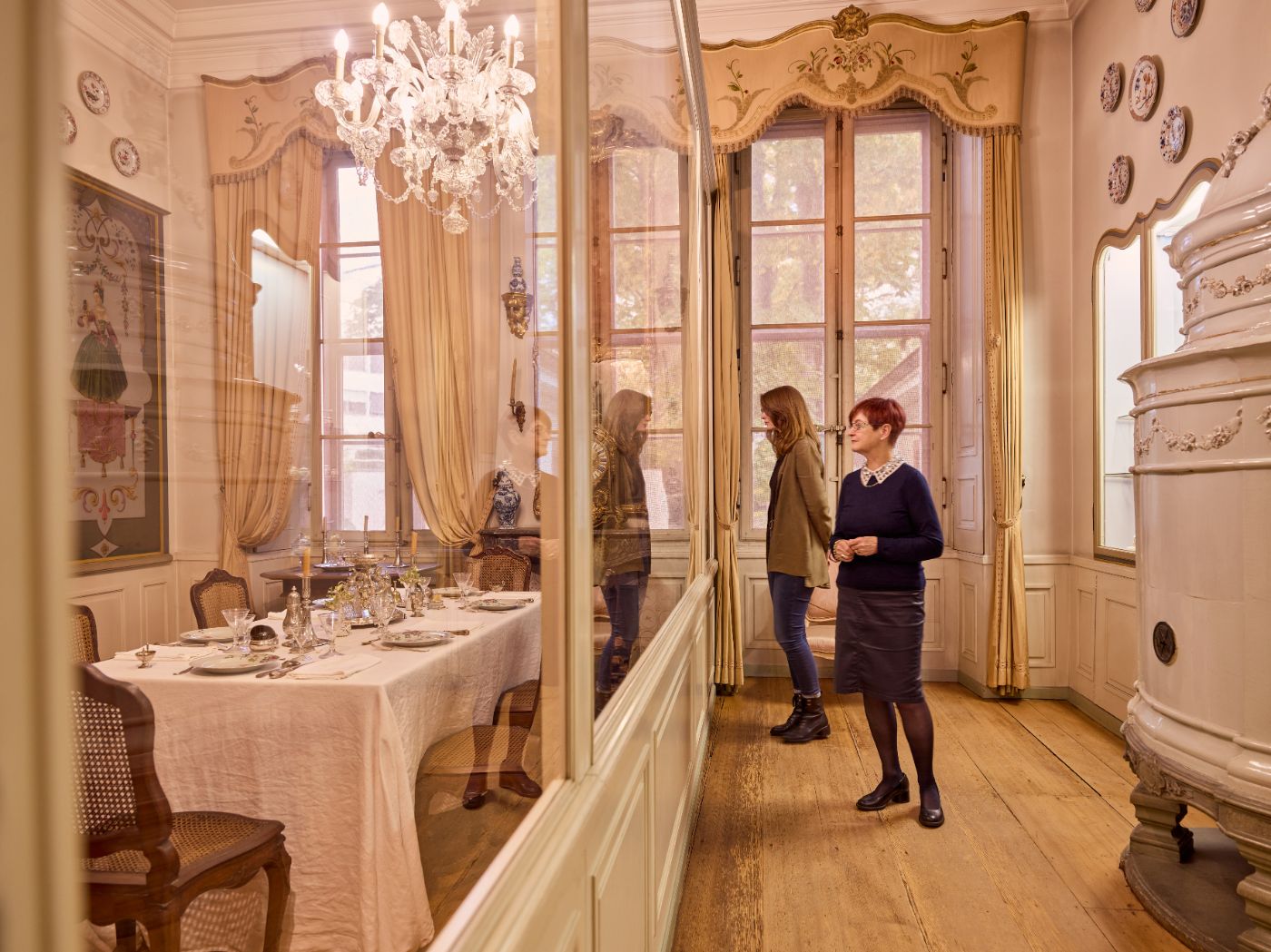 In einem prunkvollen Raum betrachten zwei Frauen durch Glas eine gedeckte Tafel
