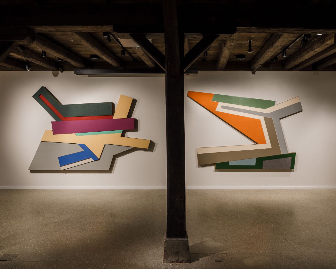 Zwei farbenfrohe geometrische Kunstwerke hängen nebeneinander an einer Holzwand.
