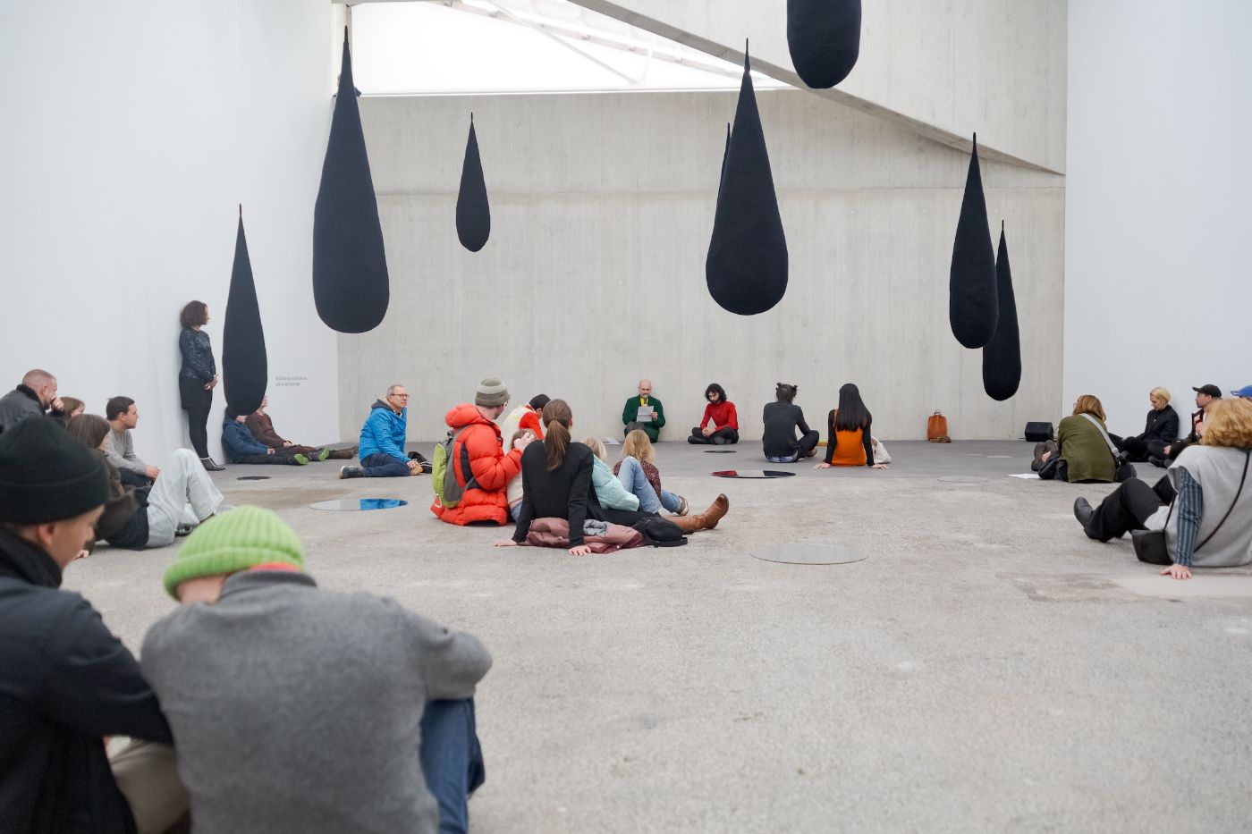 Visitors sit on a gray floor in a gallery with hanging black drop shapes