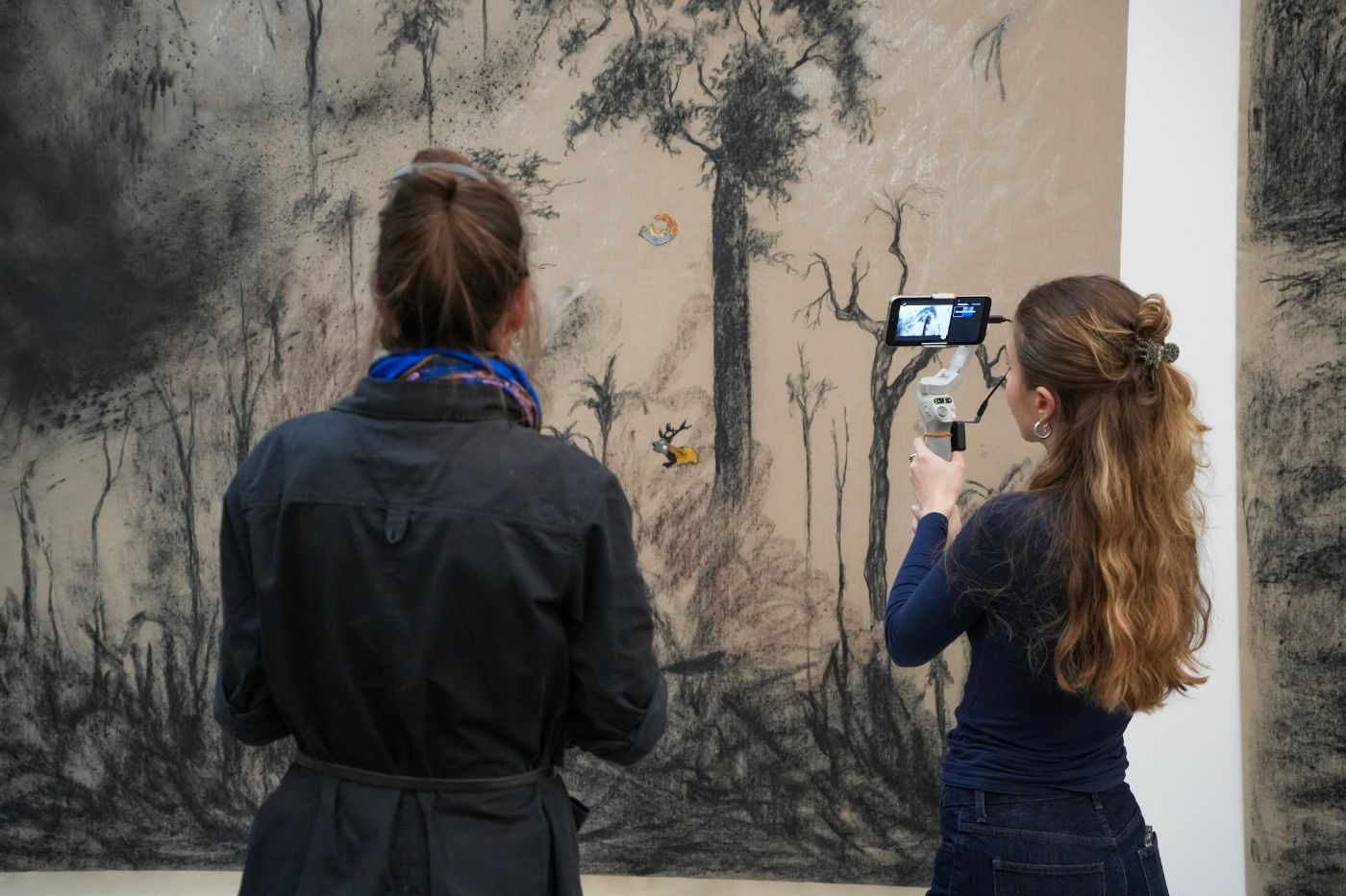 Zwei Personen betrachten ein Kunstwerk mit dunklen Bäumen, eine filmt mit einem Smartphone.