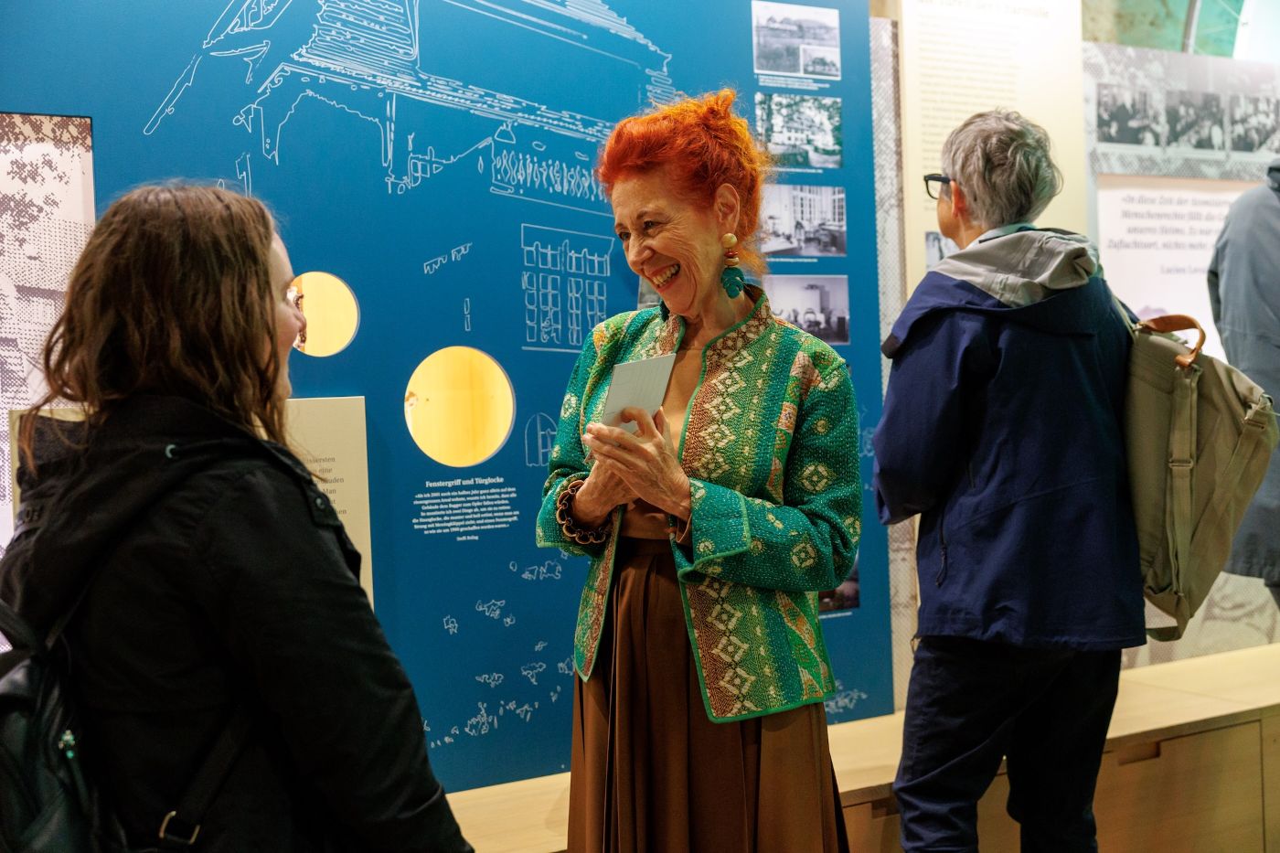 Eine Frau im bunten Outfit lächelt einer anderen Frau zu, während sie in einem Ausstellungsraum spricht.