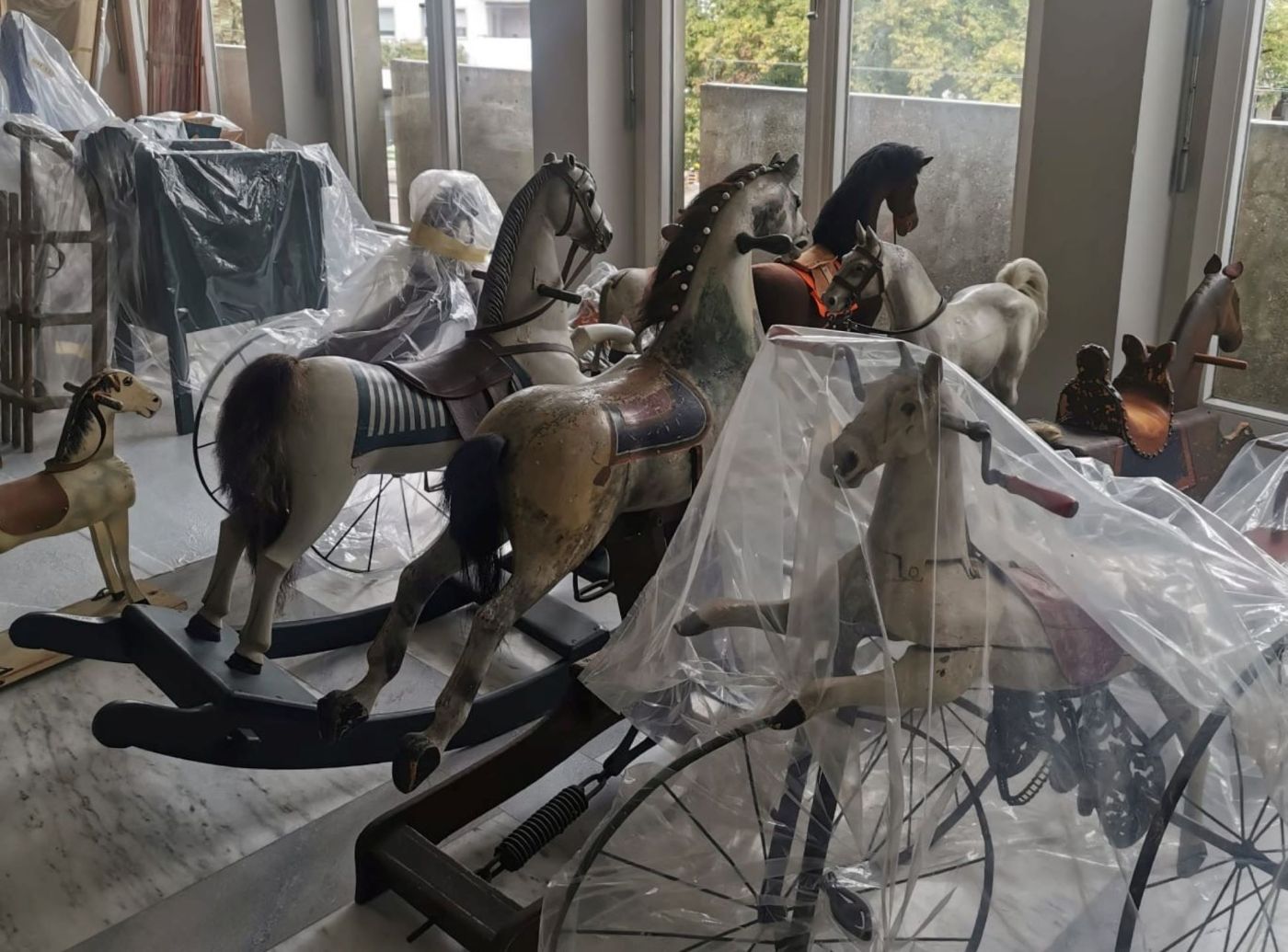 Historische Schaukelpferde stehen in einem Raum, teils von Plastikfolie bedeckt