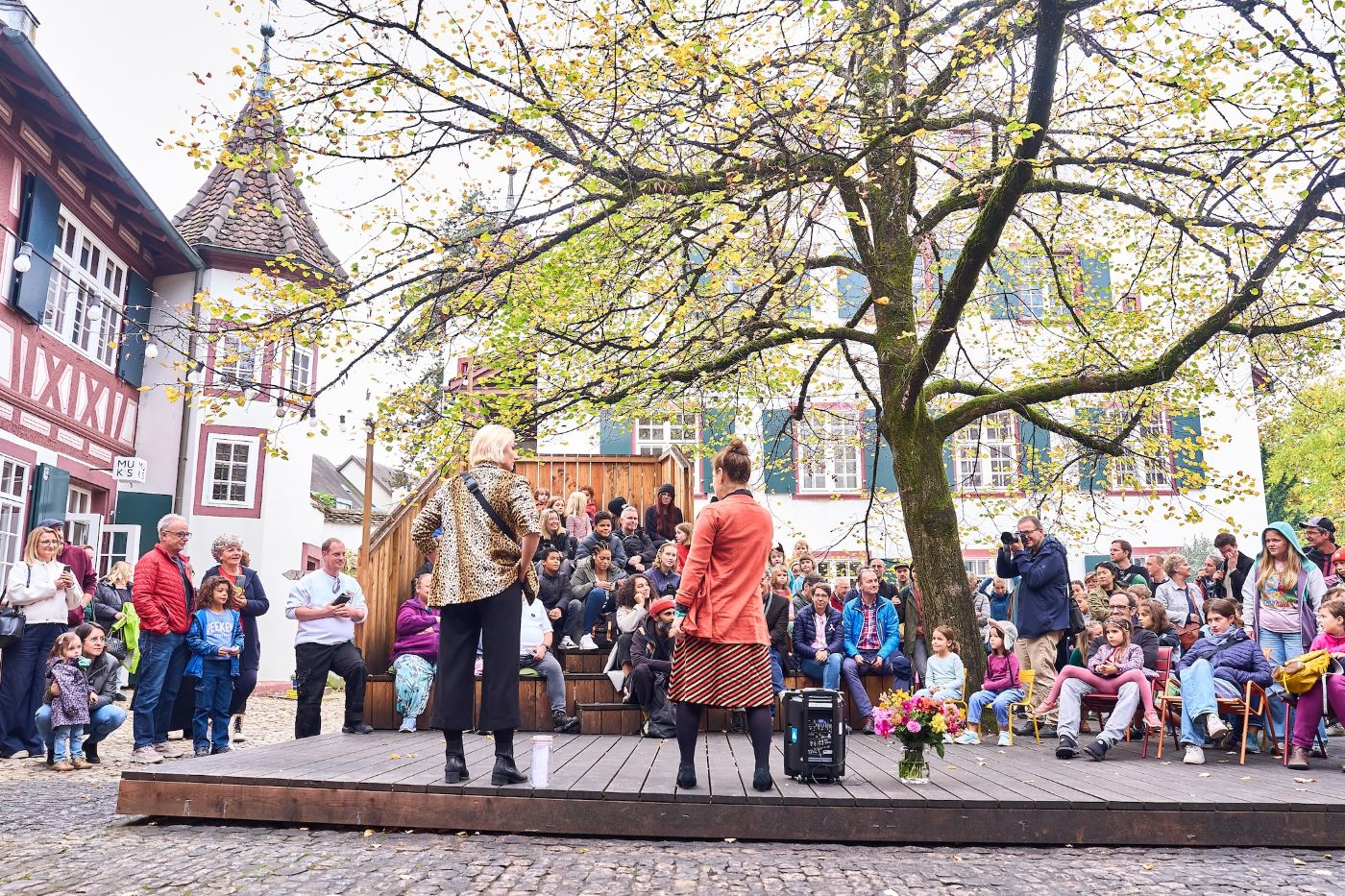 Zwei Frauen stehen auf einer Holzplattform vor einer bunt gemischten Gruppe von Zuschauern
