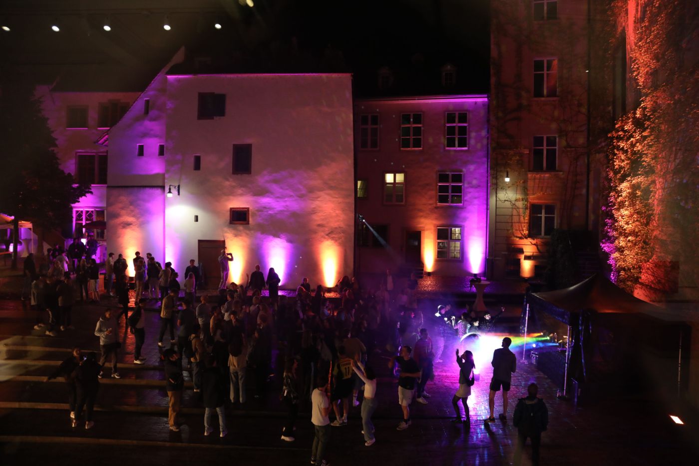 Eine Gruppe von Menschen tanzt nachts im Hof auf nassem Pflaster, während bunte Lichter leuchten