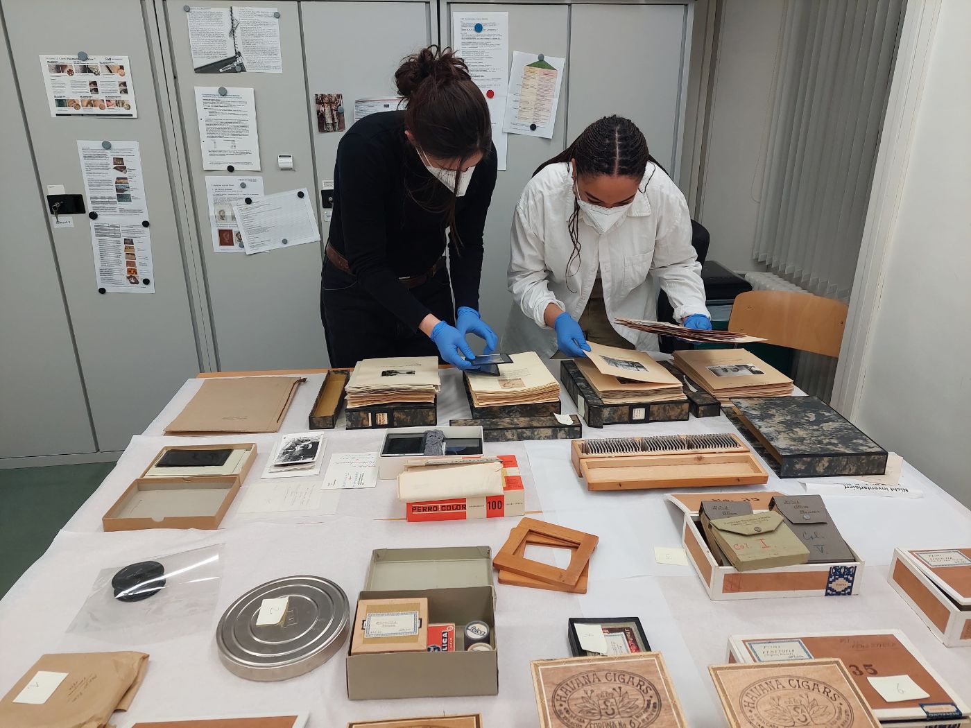 Zwei Personen mit Masken und Handschuhen sortieren historische Fotos und Dokumente an einem Tisch