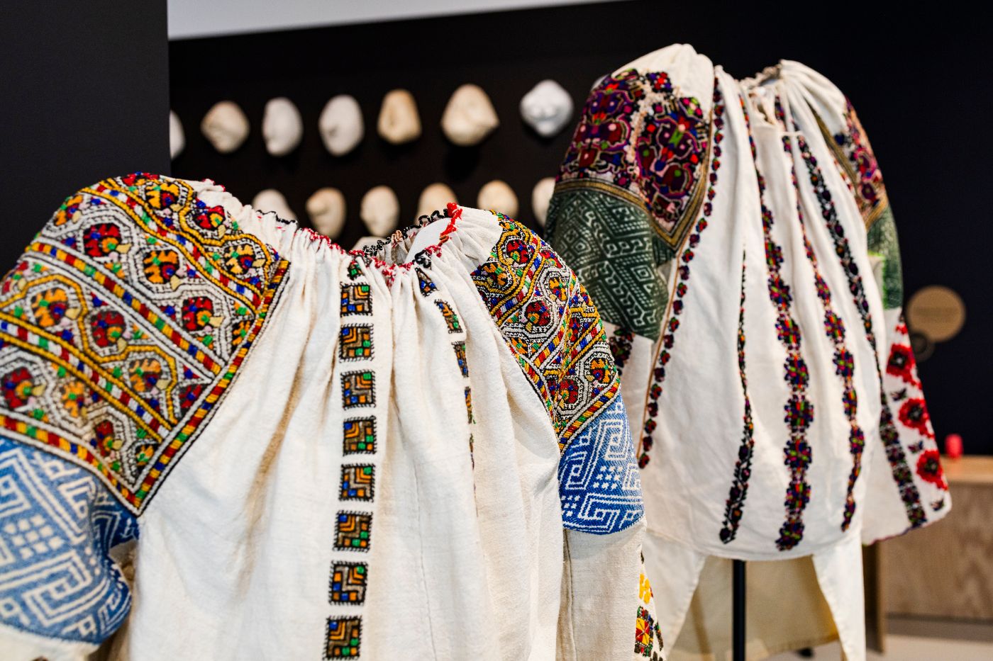 Zwei bunt bestickte weisse Blusen hängen in einem Ausstellungsraum.
