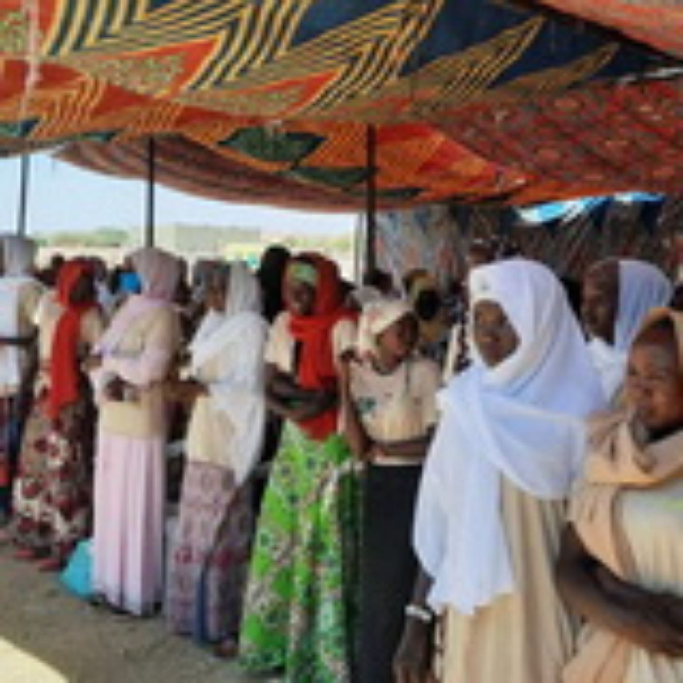 Frauen in bunten Kleidern stehen unter einem gemusterten Zelt auf sandigem Boden im Freien