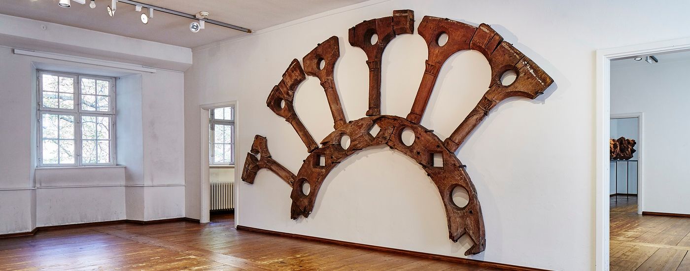 Ein grosser, fächerförmiger Holzrahmen mit kreisförmigen Öffnungen hängt an einer weissen Museumswand