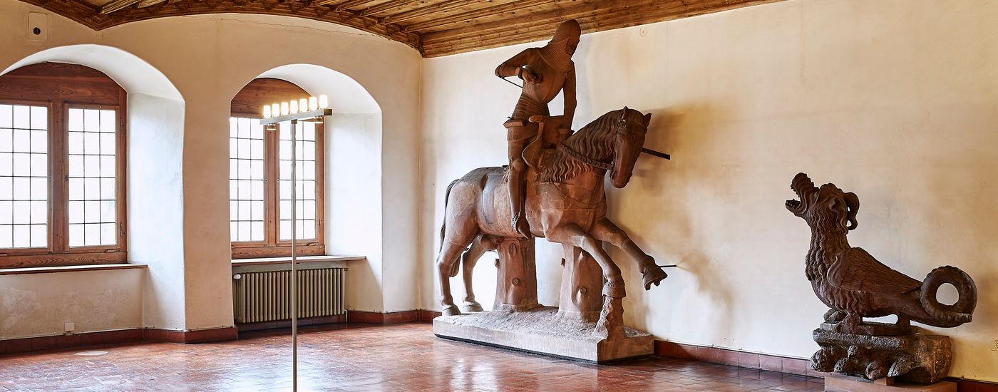 Ein Ritter aus Stein reitet auf einem Pferd neben einem Fabelwesen in einem lichtdurchfluteten Raum