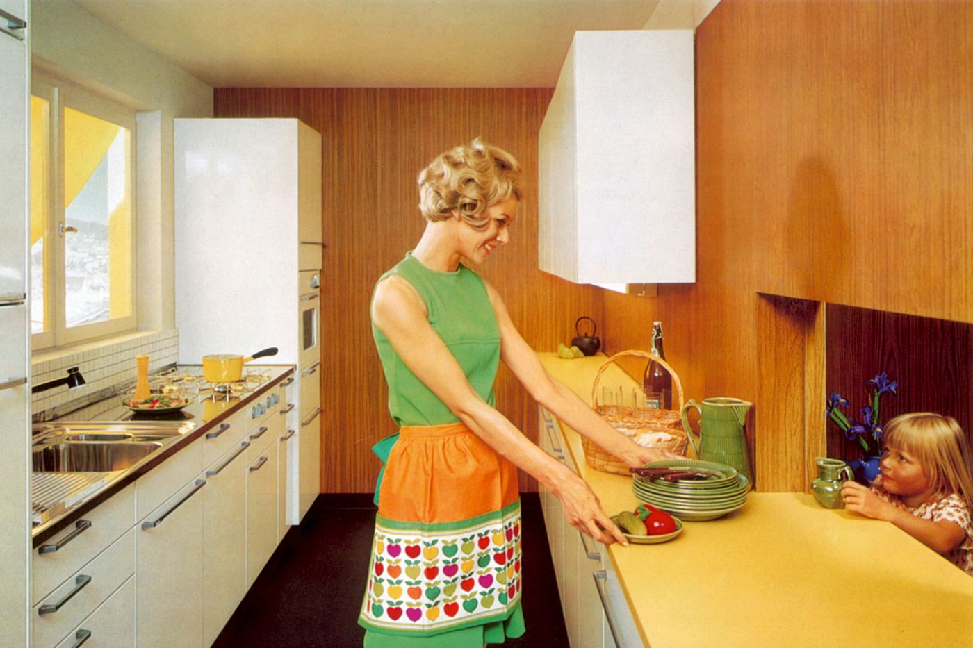 Eine Frau bereitet in einer Küche im Stil der 1960er Jahre ein Gericht zu und reicht es einem Kind über die Theke.