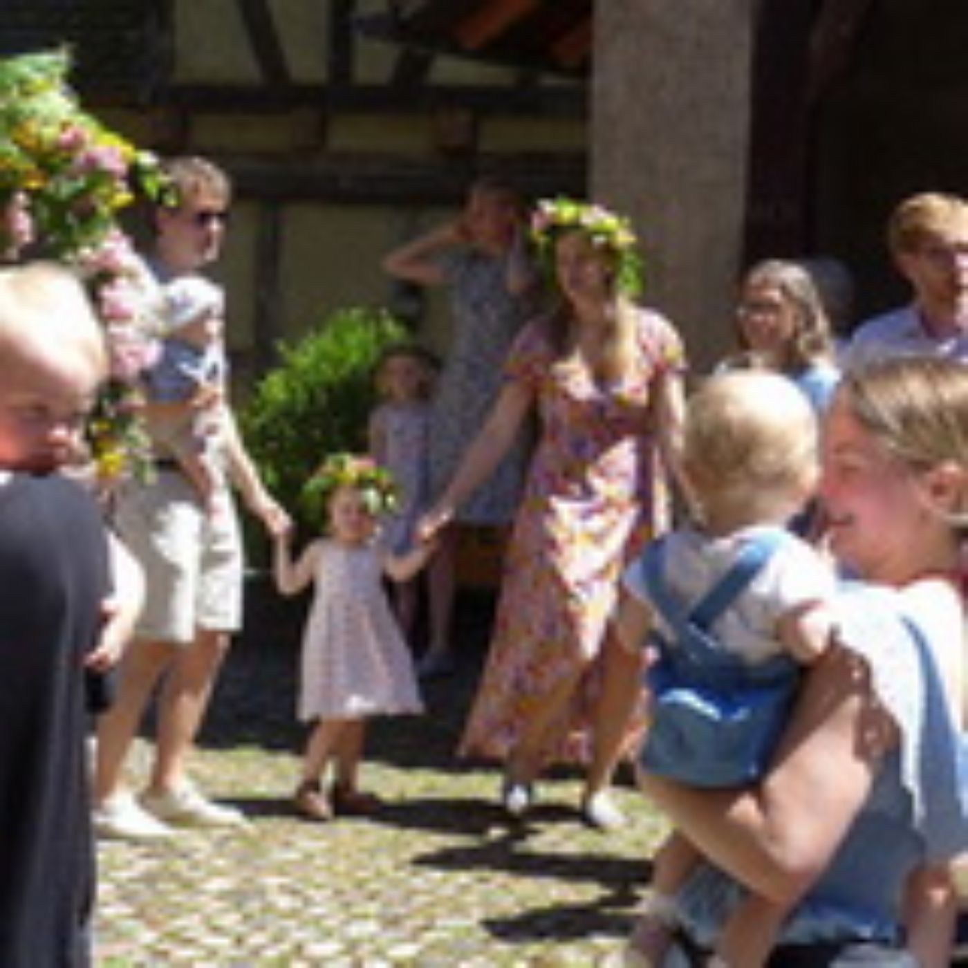 Eine Gruppe von Menschen tanzt draussen, Blumenkränze auf den Köpfen und Kinder in den Armen.