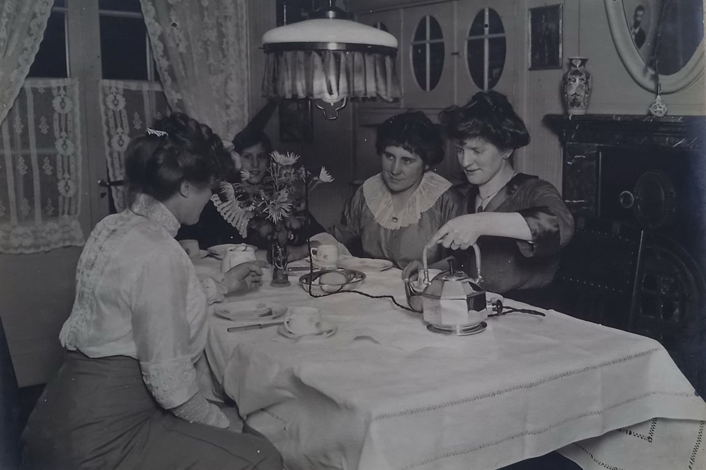 Vier Frauen sitzen bei einer Tasse Tee zusammen an einem Tisch, eine Frau schenkt aus einer elektrischen Teekanne ein.