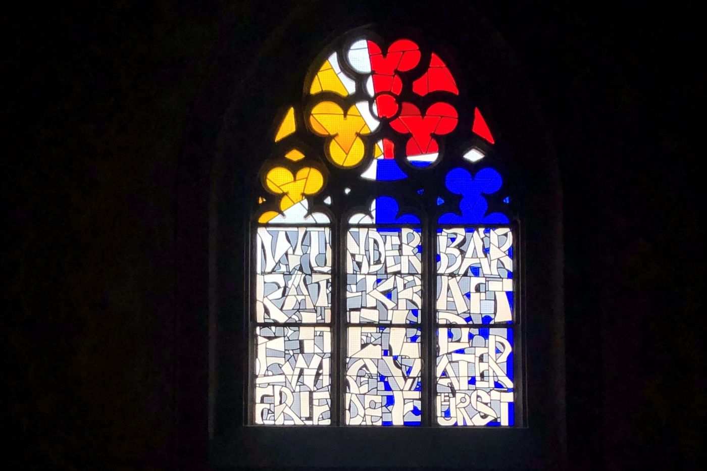 Ein buntes Glasfenster zeigt abstrakte Formen in Gelb, Rot und Blau sowie lesbare Wörter