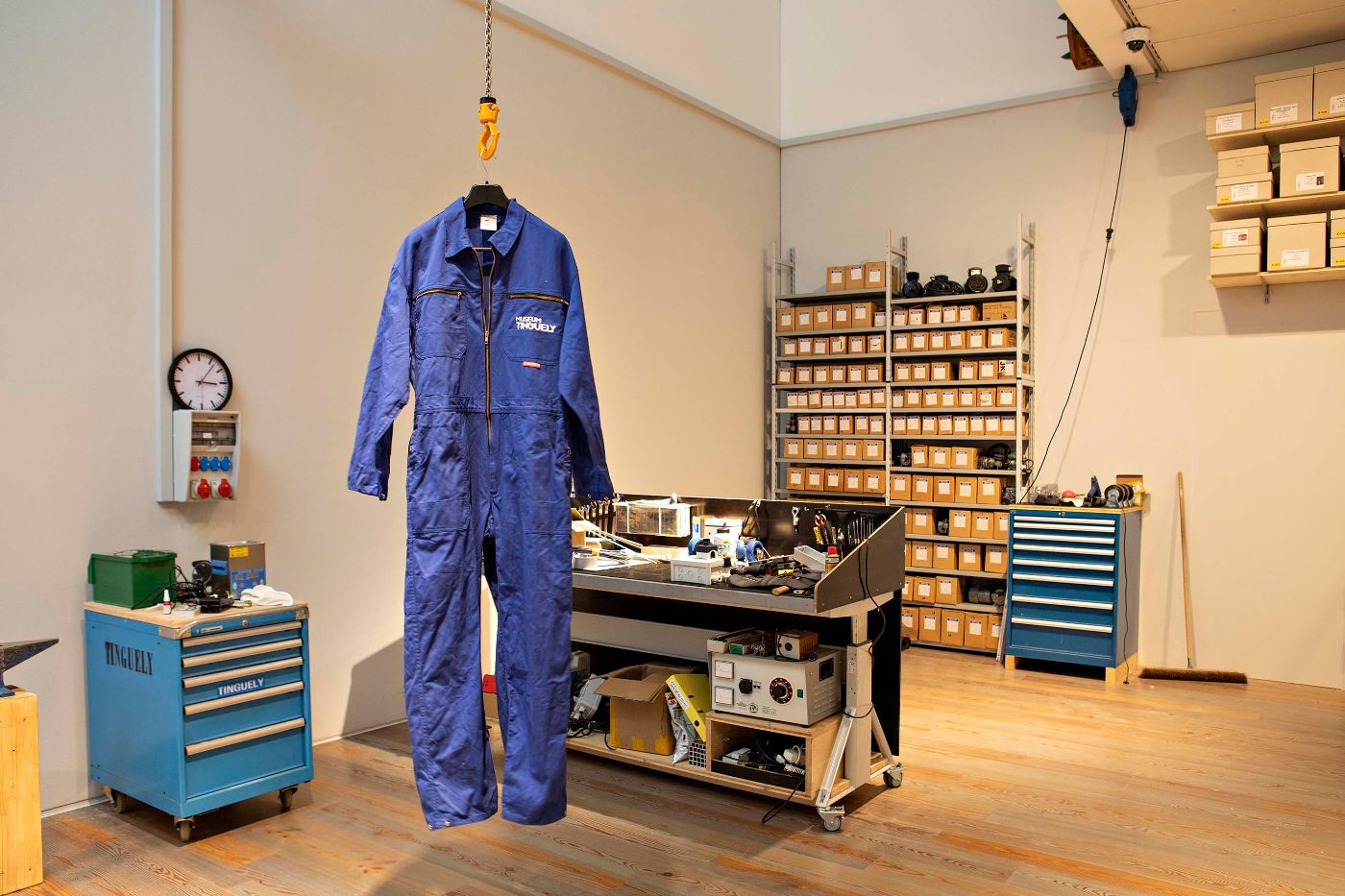 Ein blauer Overall hängt in einer Werkstatt mit Regalen voller Kisten und einem Holzboden.