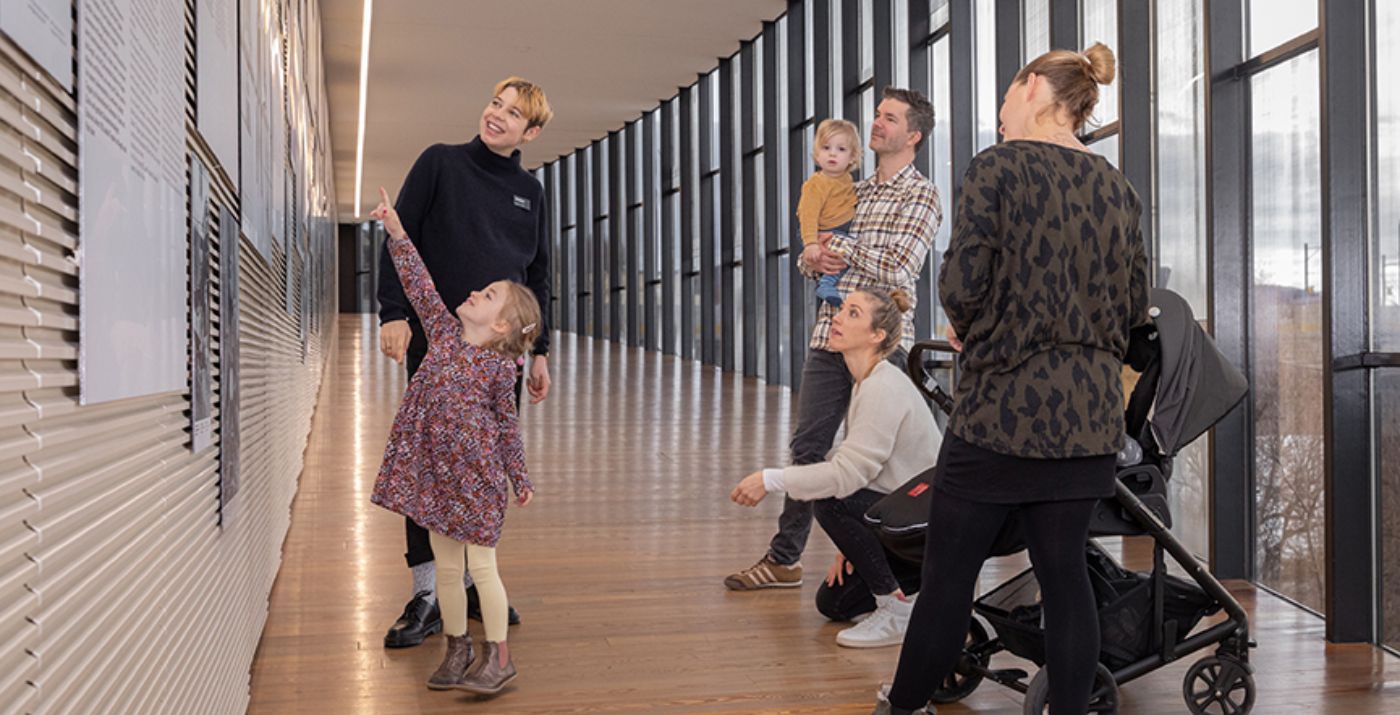 Auf einem hölzernen Museumsflur betrachten Erwachsene und Kinder gemeinsam eine Wandinstallation