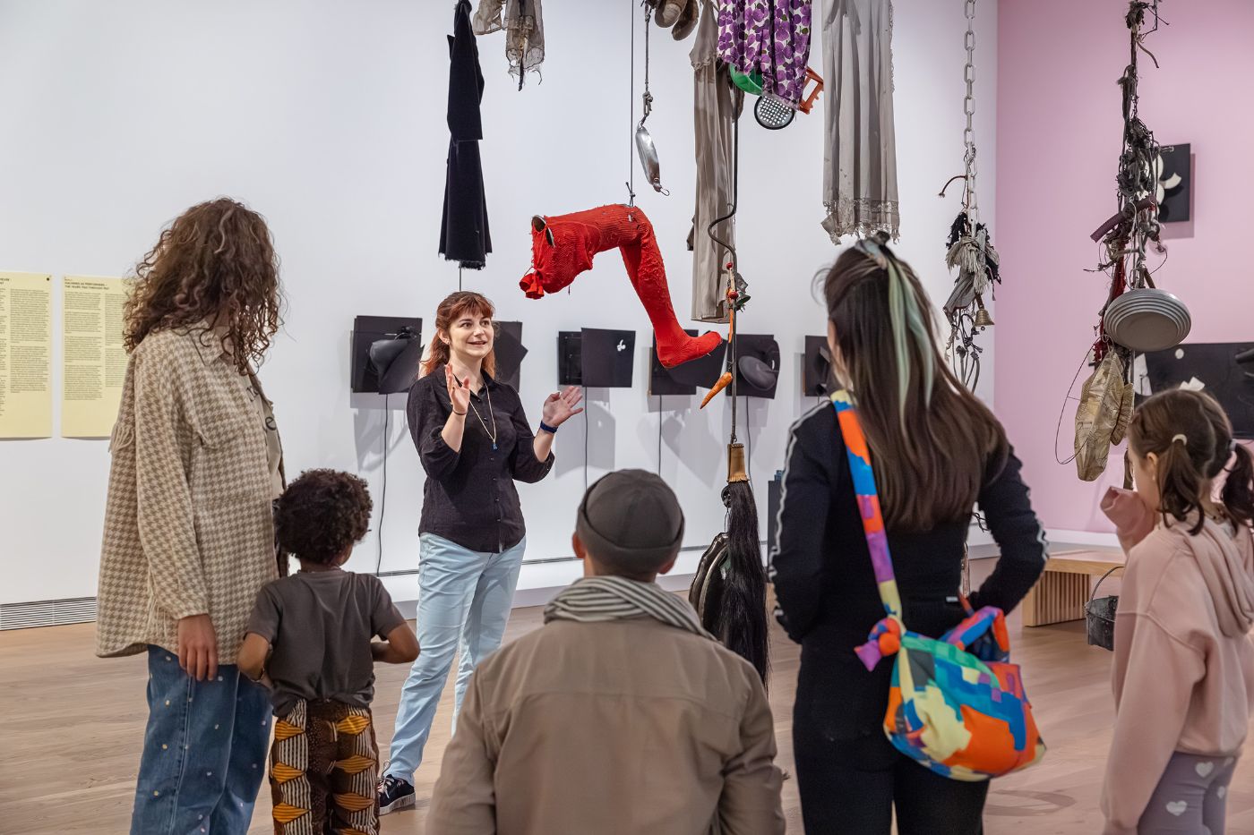 Eine Museumsführerin erklärt Kindern und Erwachsenen eine Installation mit hängenden Kleidungsstücken und Gegenständen an einer weißen Wand. 