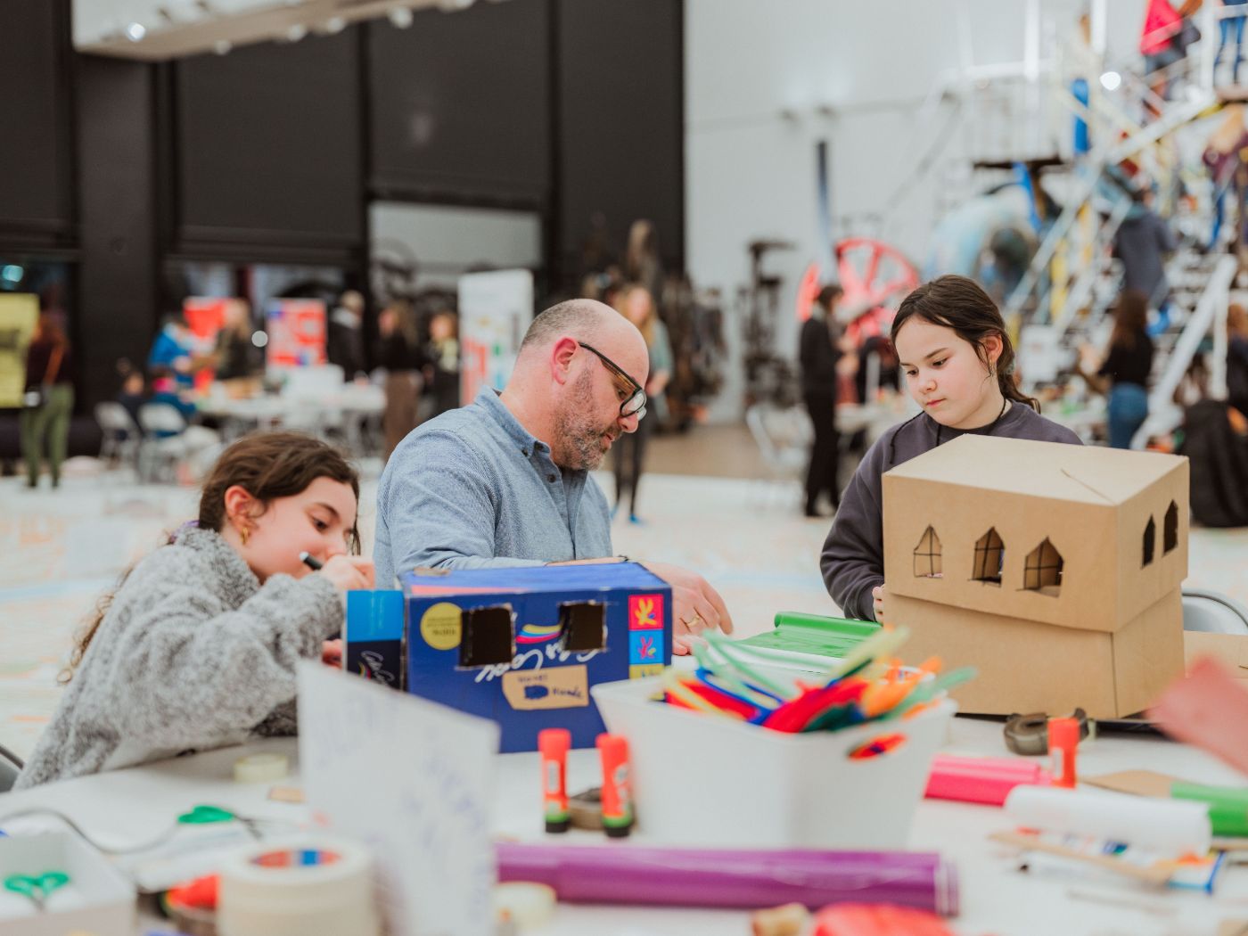 Ein Vater und zwei Kinder basteln konzentriert an einem Tisch in einem museumähnlichen Raum