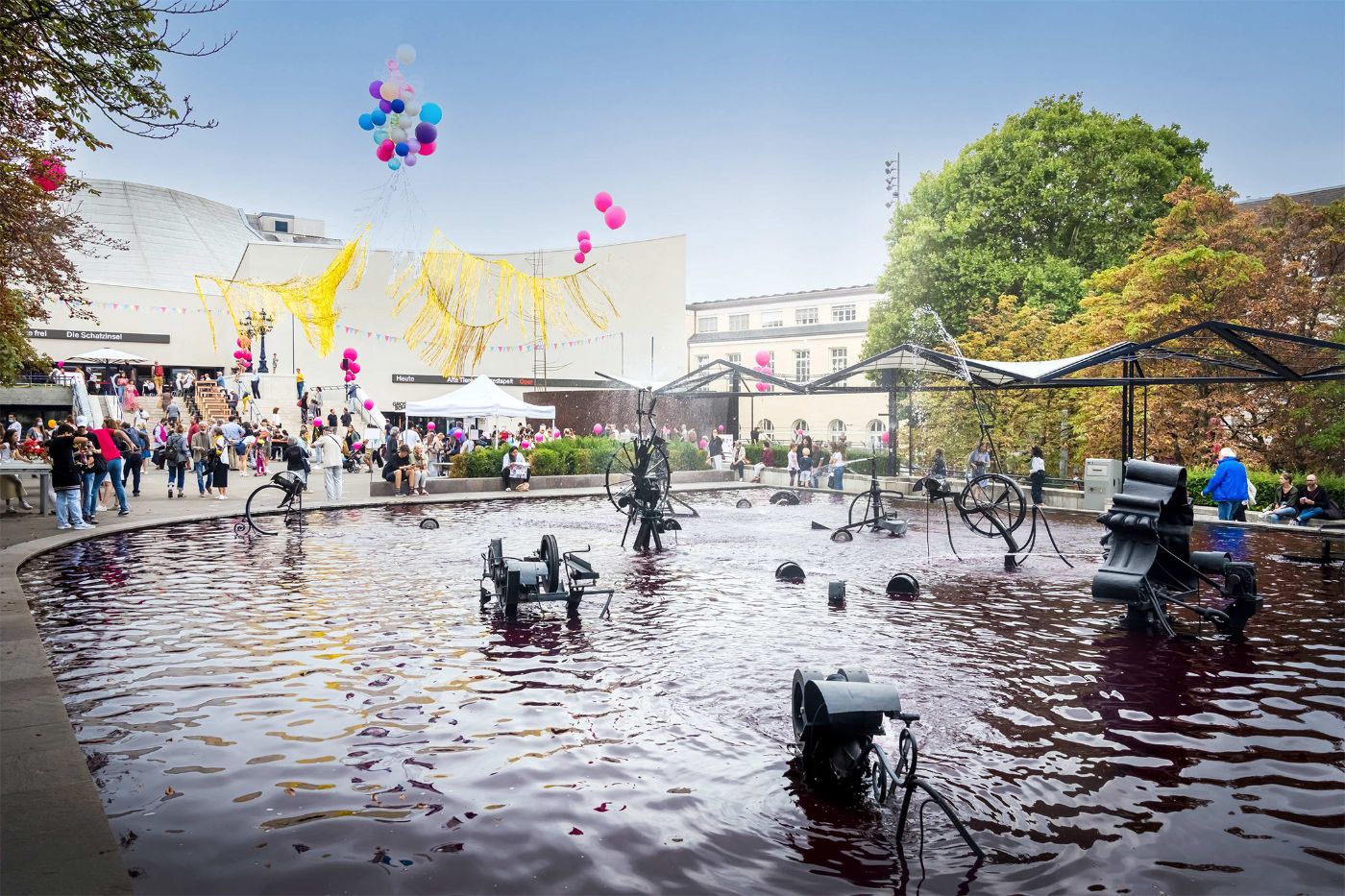 Menschen geniessen ein Fest um einen Brunnen mit bunten Ballons, während Skulpturen spritzen