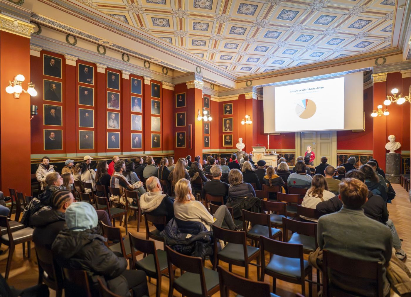 Publikum lauscht Vortrag in historischem Saal mit Porträts und Deckendekorationen. Präsentation projiziert.