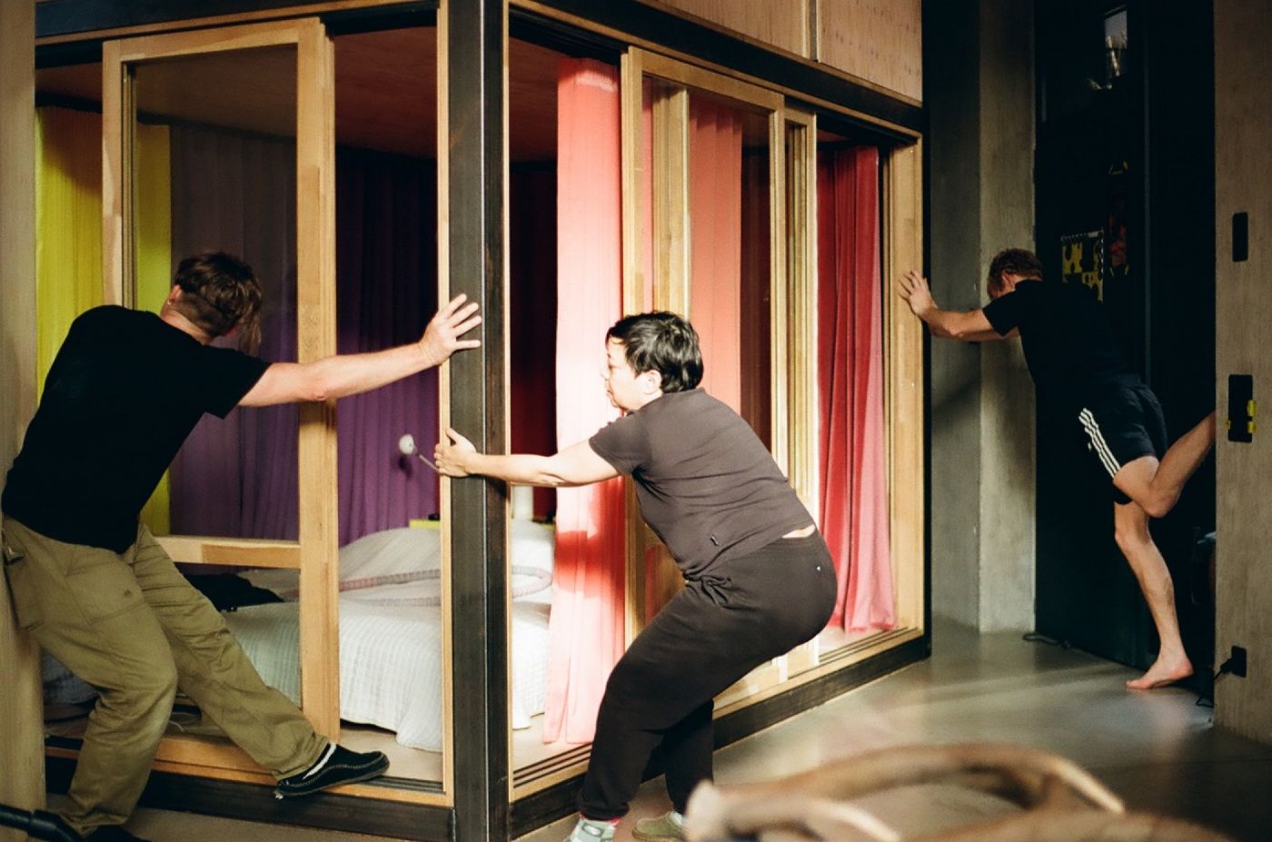 Drei Personen bewegen eine verschiebbare Holzwand in einem Raum mit bunten Vorhängen