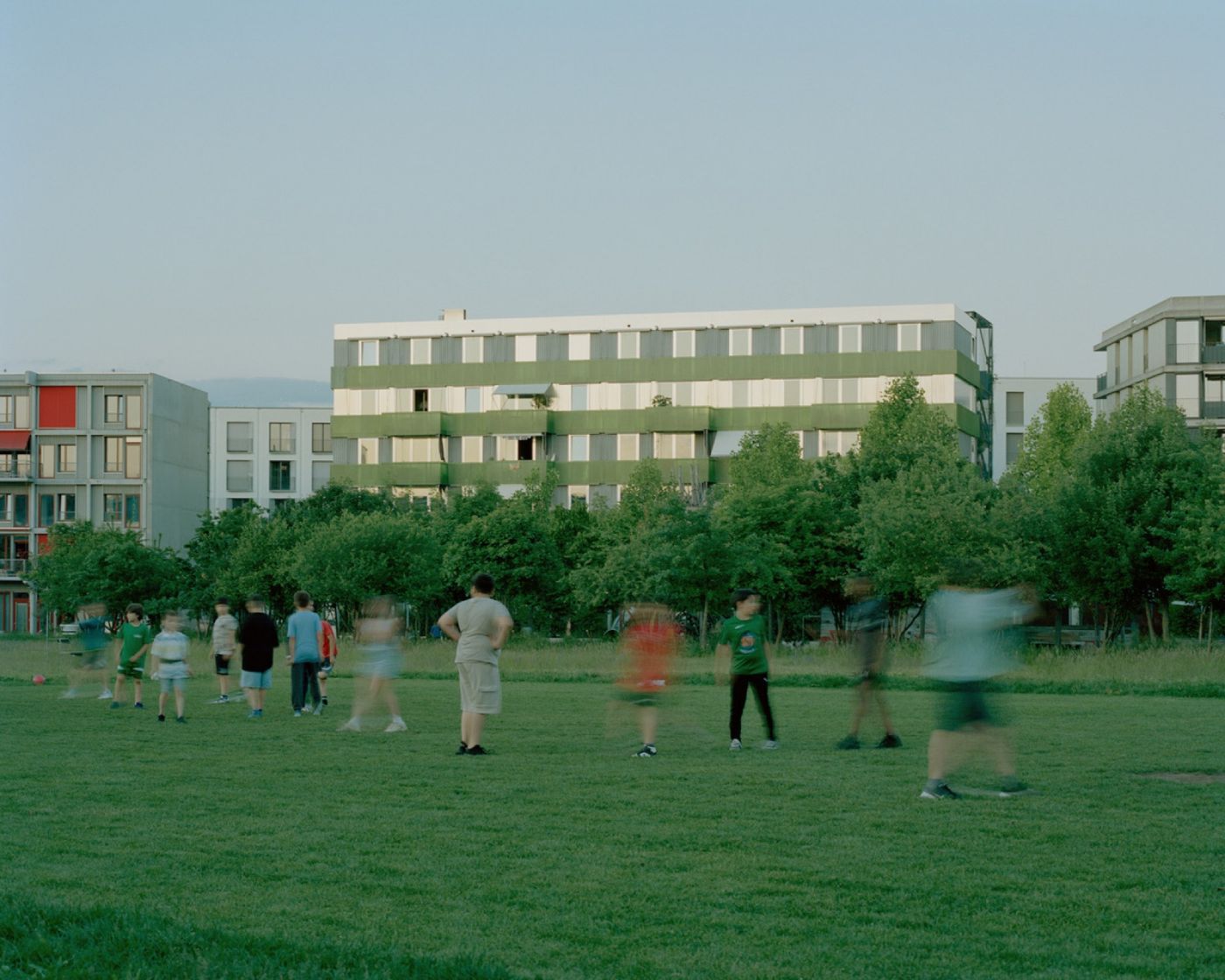Eine Gruppe unscharfer Kinder spielt auf einer Wiese vor modernen Wohngebäuden am Abend